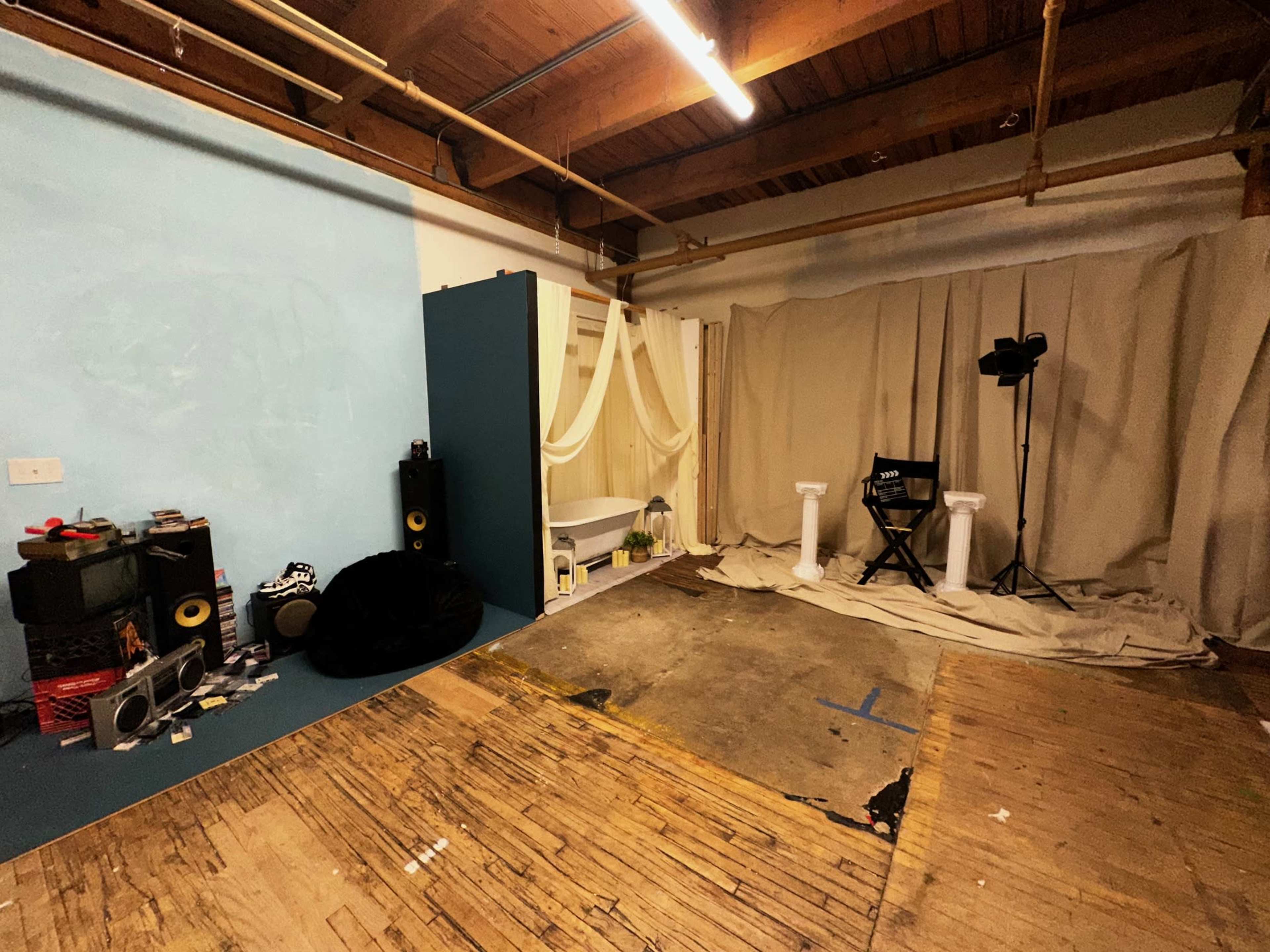 The image shows an unfinished studio space with a light blue wall, a few items including speakers and a black bean bag, and a backdrop draped on one side.