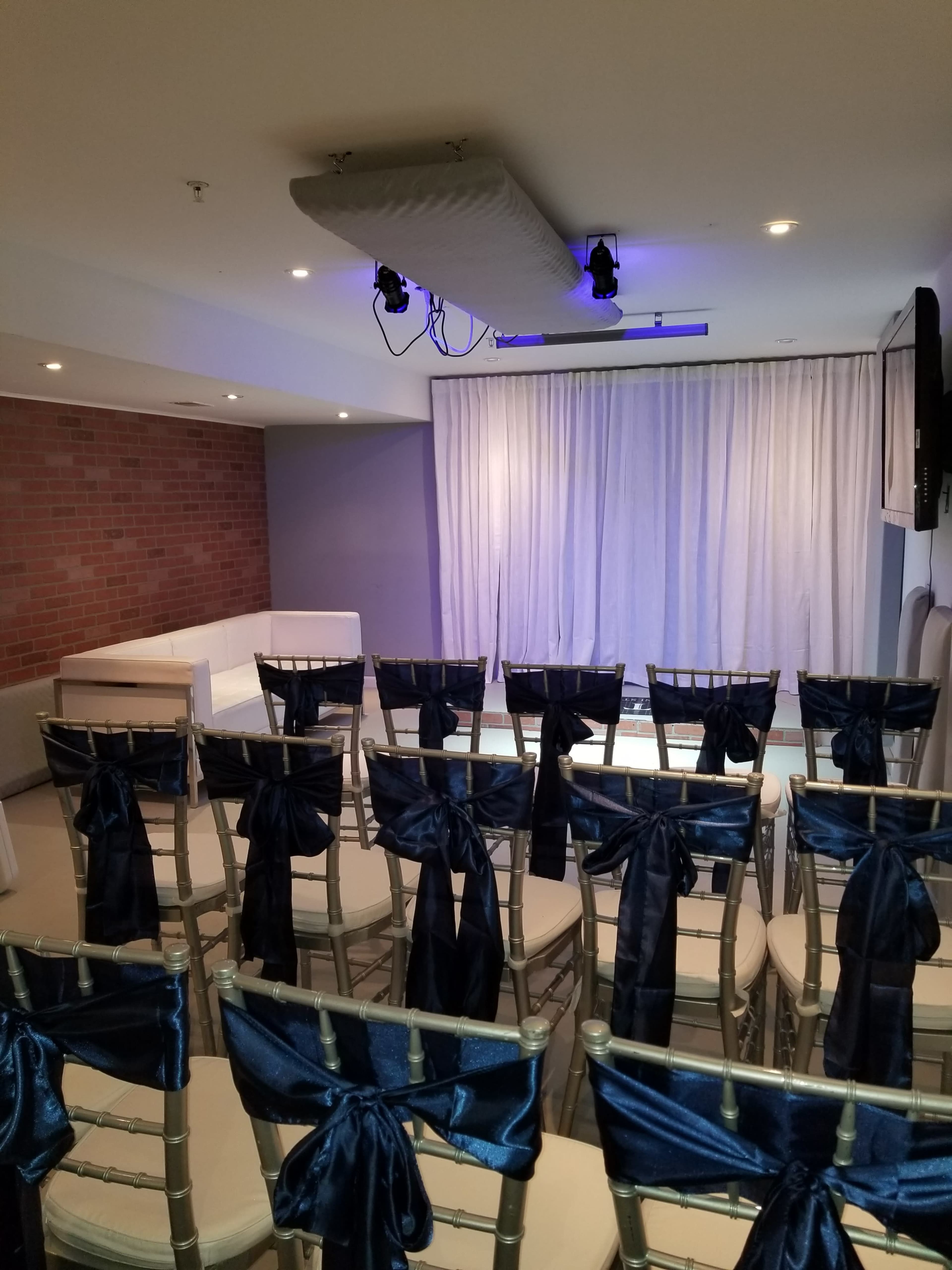 A room set up for an event with rows of chairs draped in navy blue ribbons facing a stage with a white curtain.