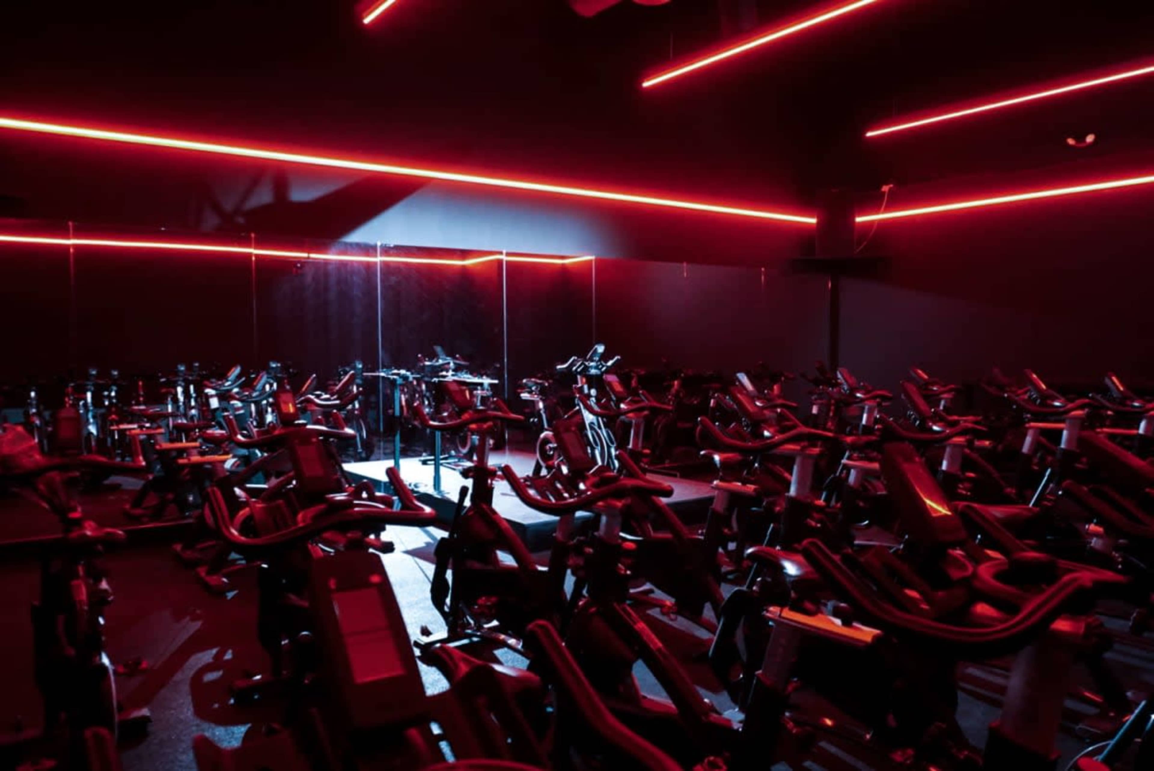 A dimly lit indoor cycling studio features rows of stationary bikes illuminated by red neon lights.