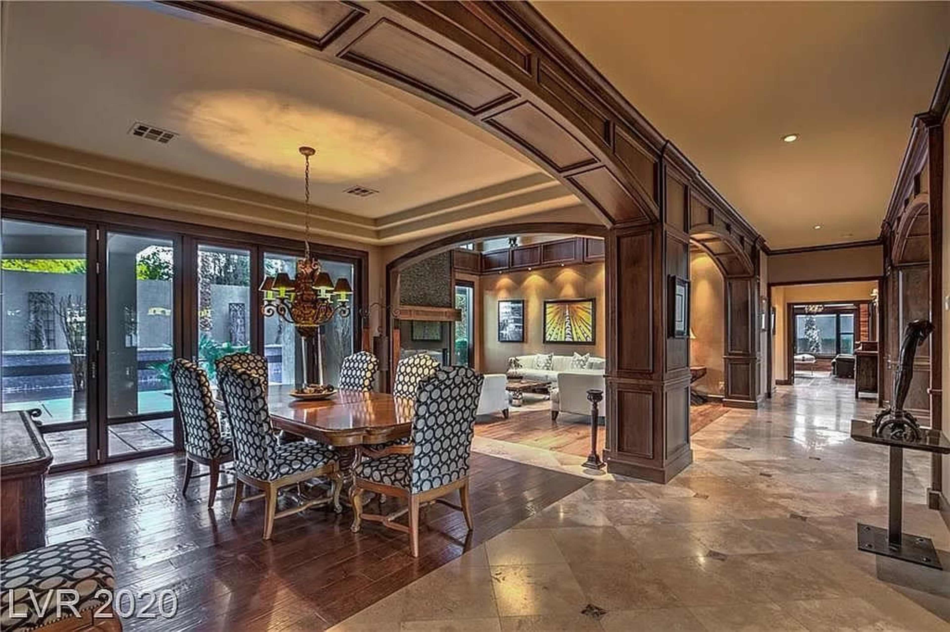 The image shows a spacious dining area featuring a wooden table with patterned chairs, surrounded by large windows with views of an outdoor space.