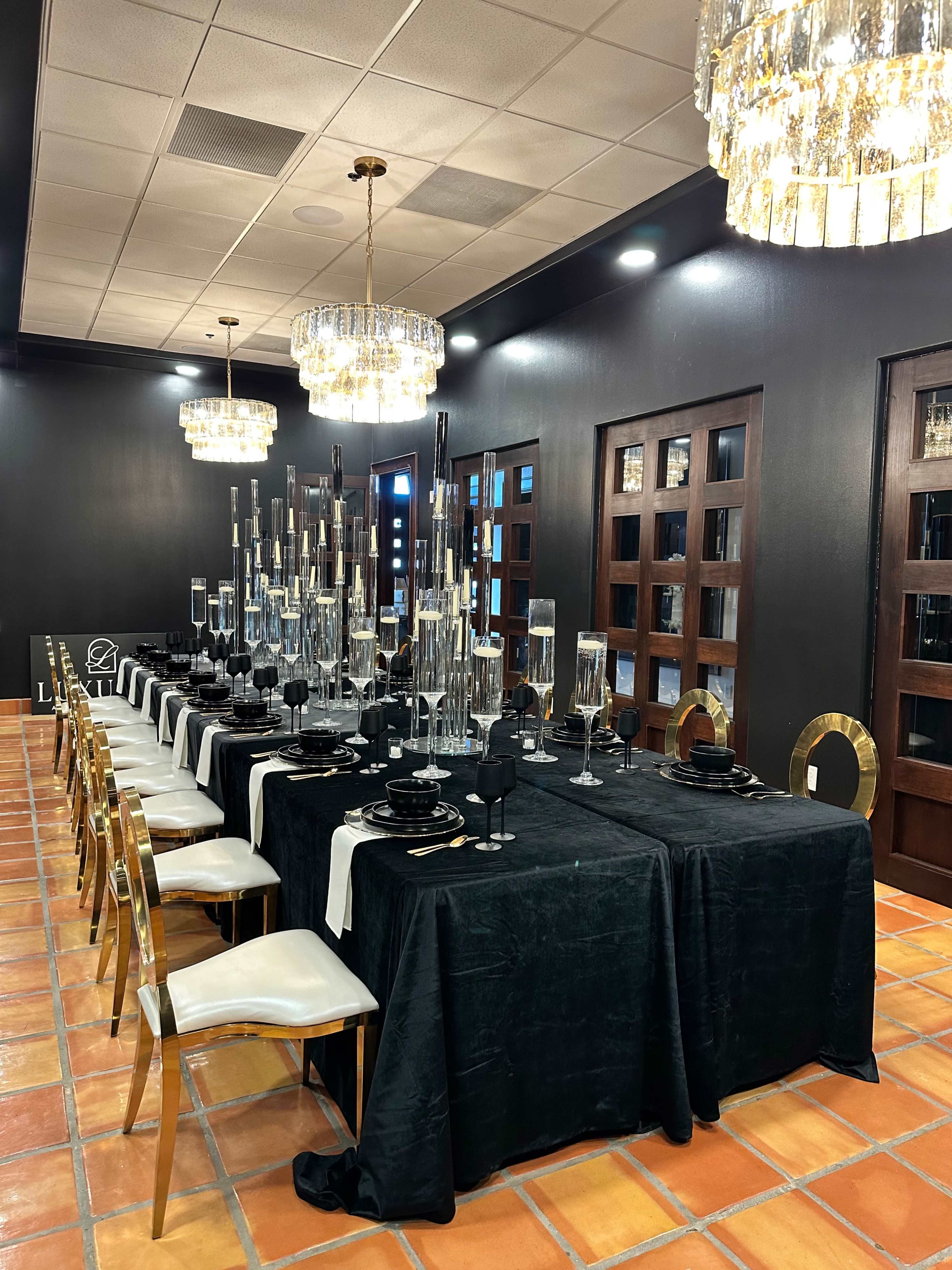 A long dining table is elegantly set with black tablecloths, glassware, and candles, surrounded by gold chairs under modern chandeliers in a dimly lit room.