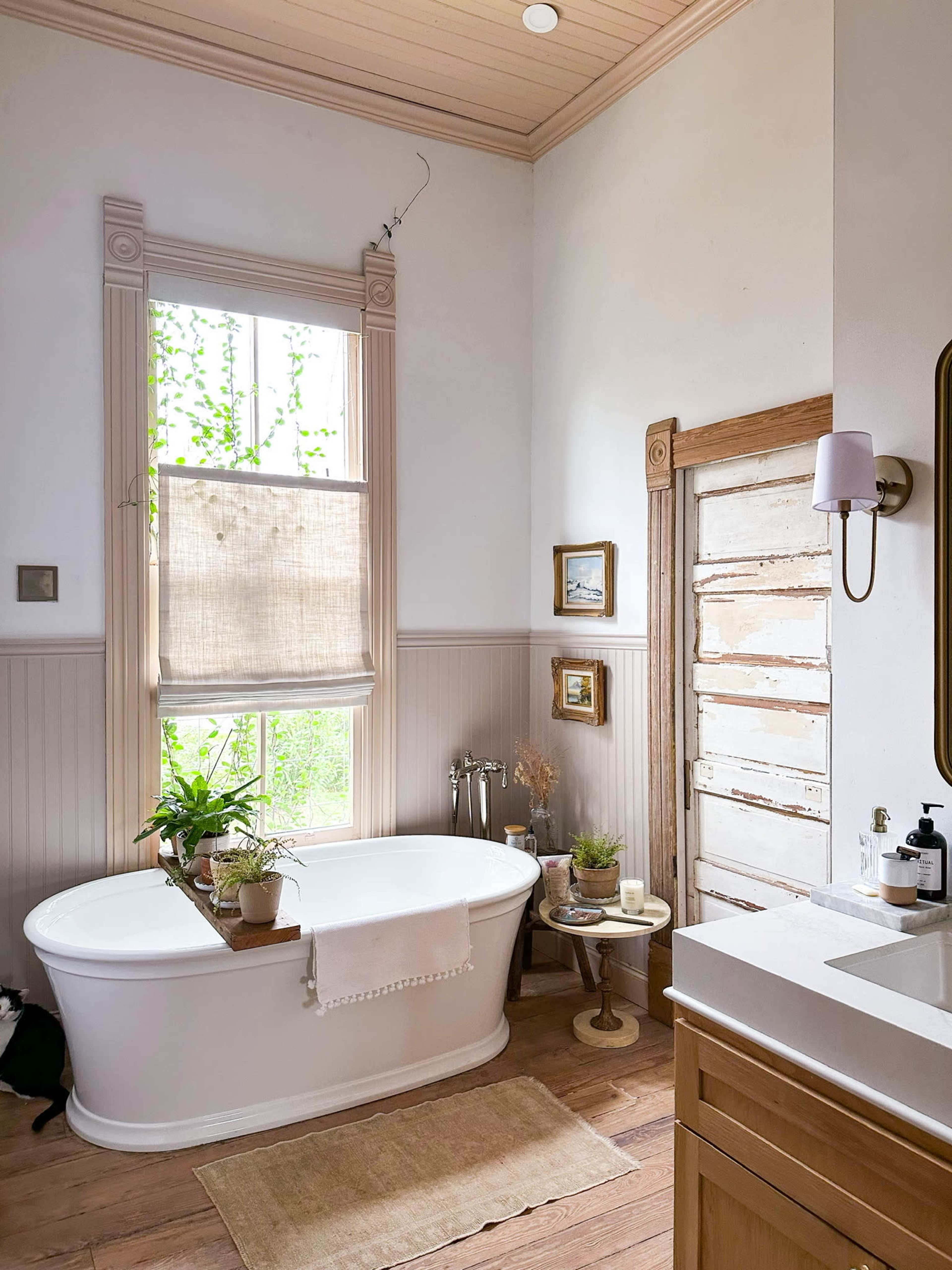A bathroom featuring a freestanding bathtub beside a window with natural light, adorned with potted plants and framed artwork on the walls.