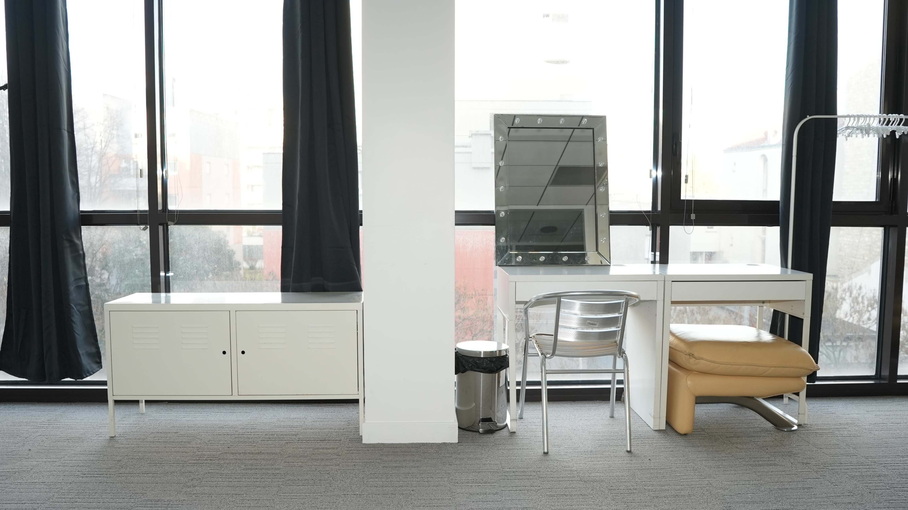 The image shows a modern interior with a white desk and a mirrored vanity set against large windows, with a storage unit and a chair nearby.