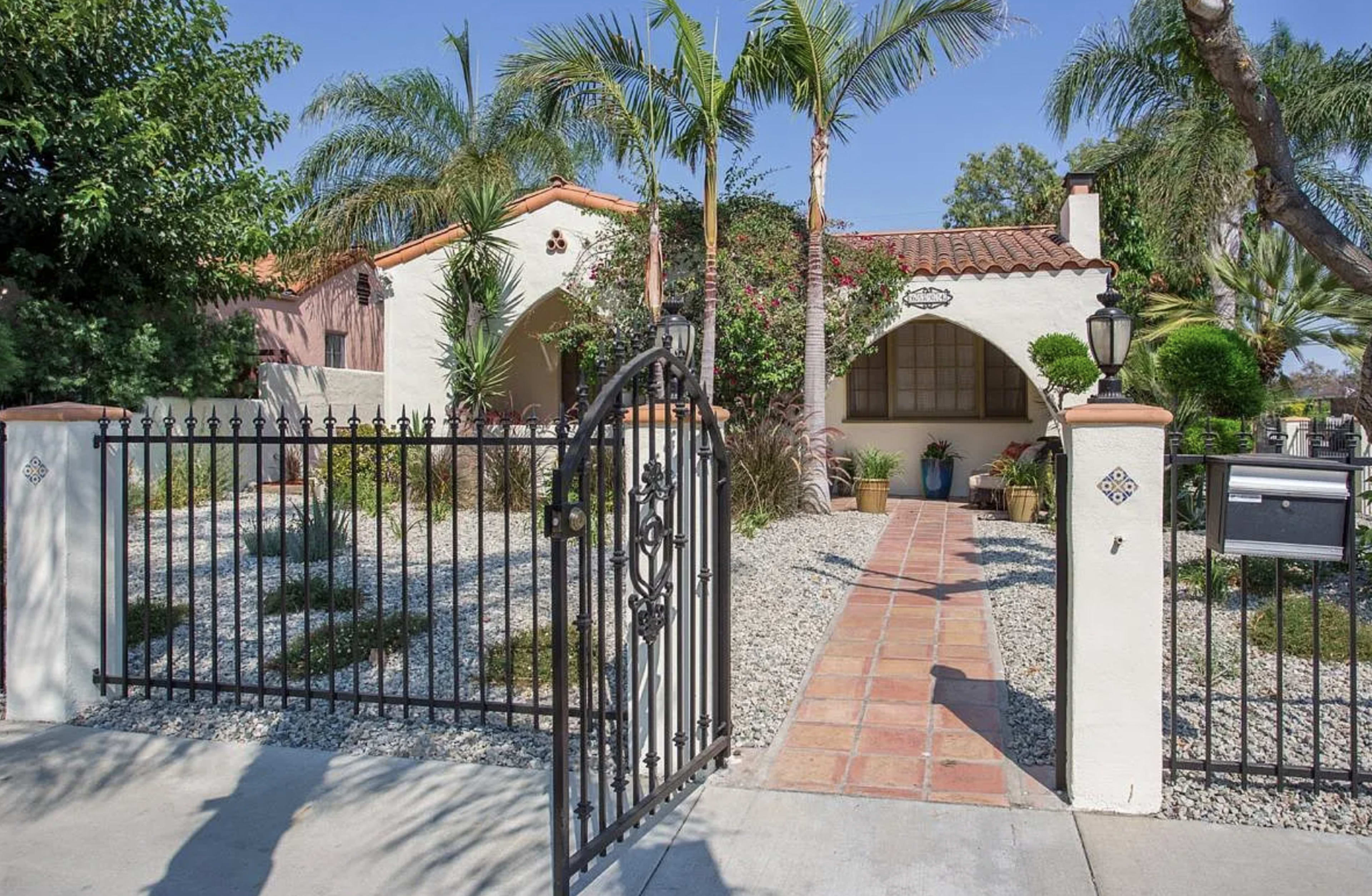 The image shows a Mediterranean-style house surrounded by lush greenery and a gravel landscape, with a decorative iron gate leading to a tiled pathway.
