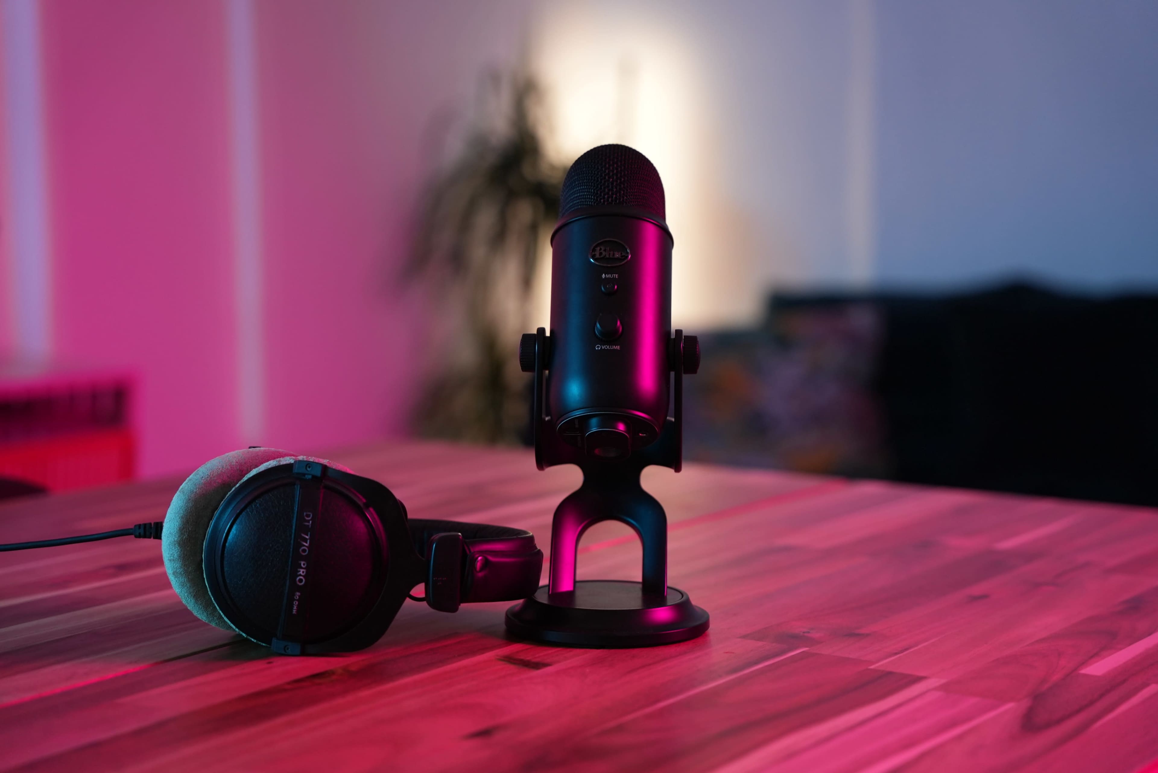 A black microphone and a pair of headphones are positioned on a wooden table, with soft, colored lighting in the background.