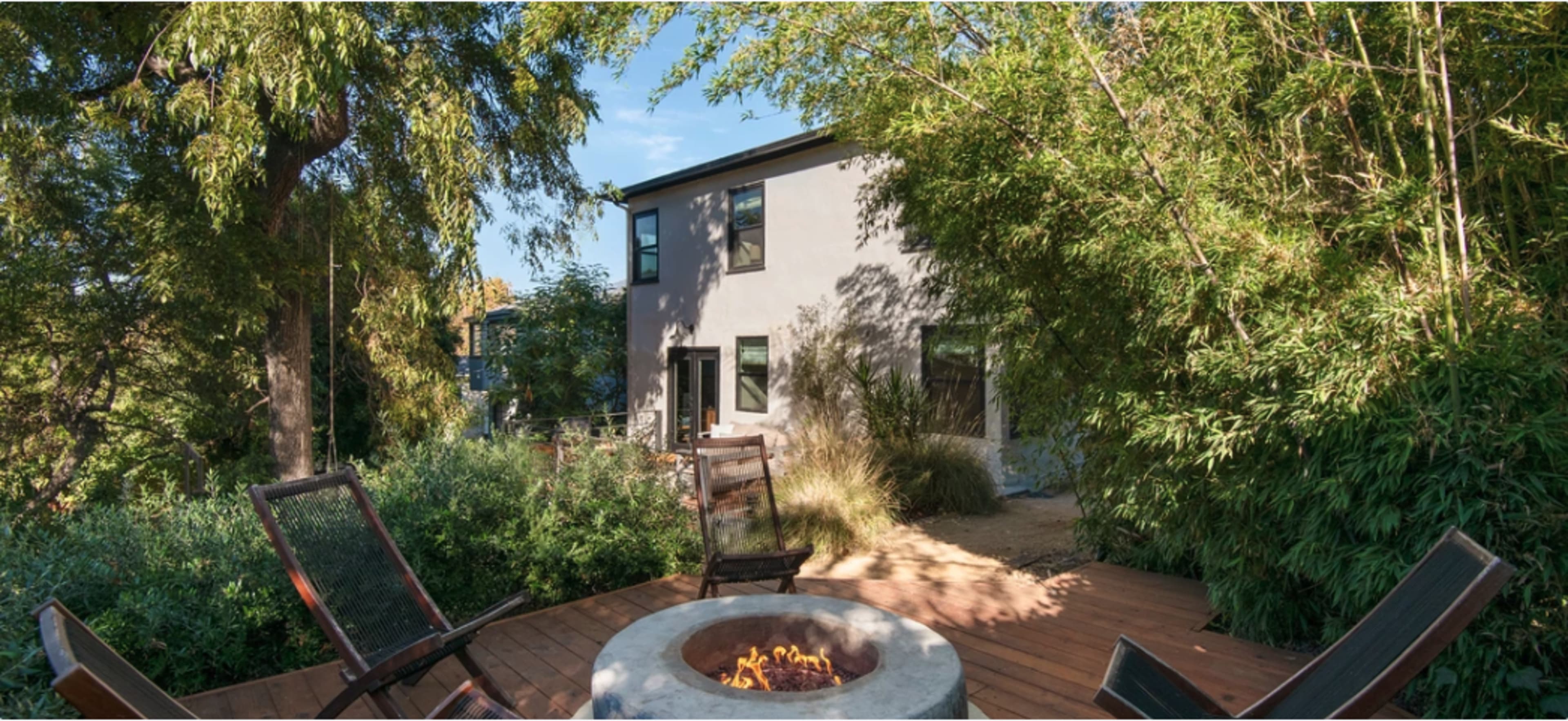 A modern house with large windows is surrounded by greenery and features a wooden deck with a fire pit and chairs in the foreground.