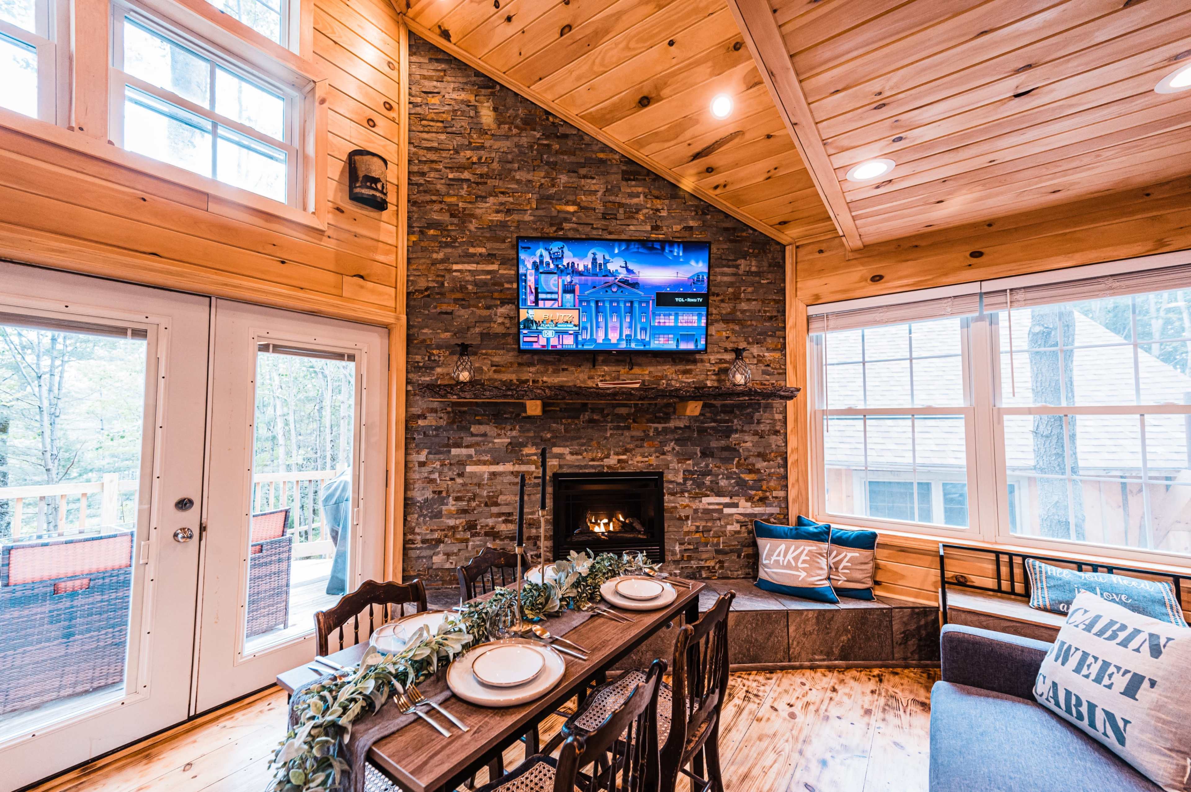 A rustic dining area with a wooden table set for a meal, a stone fireplace, and a television mounted above it, all within a cabin-like interior featuring wood paneling and large windows.