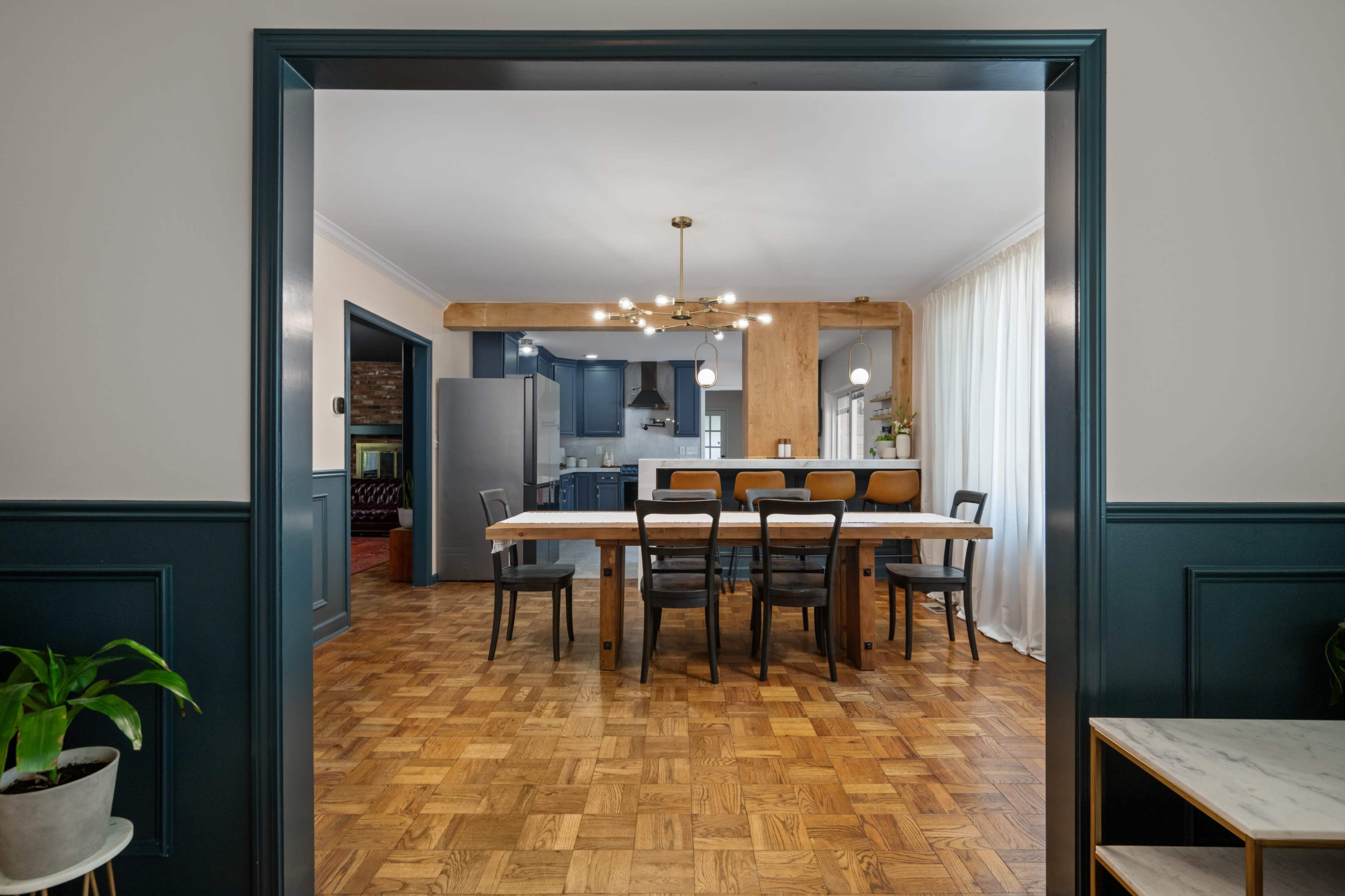 A modern dining area features a long table with several chairs, surrounded by a sleek kitchen and large windows.