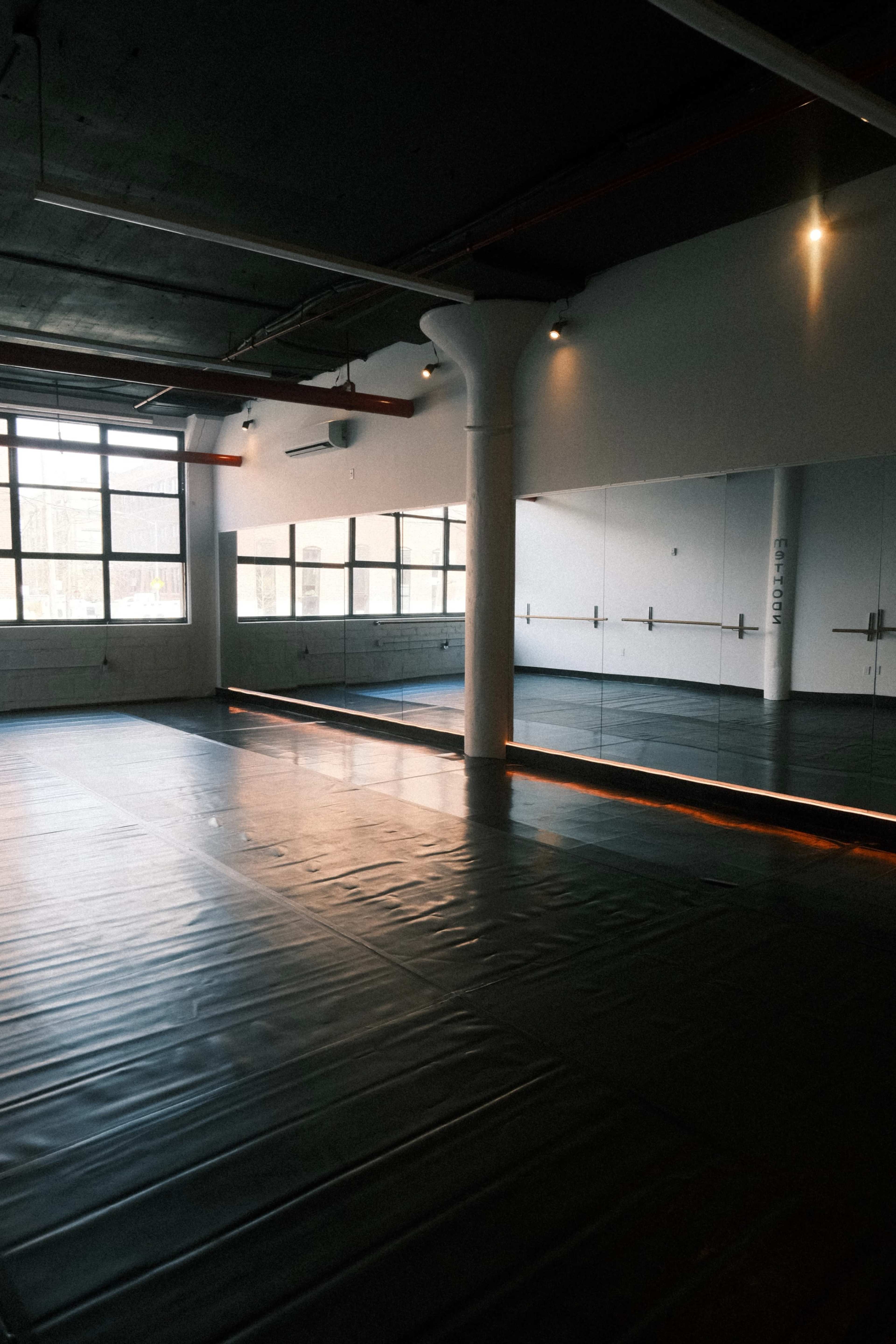 The image shows a spacious empty room with a black mat flooring and large mirrors along one wall, illuminated by natural light from large windows.