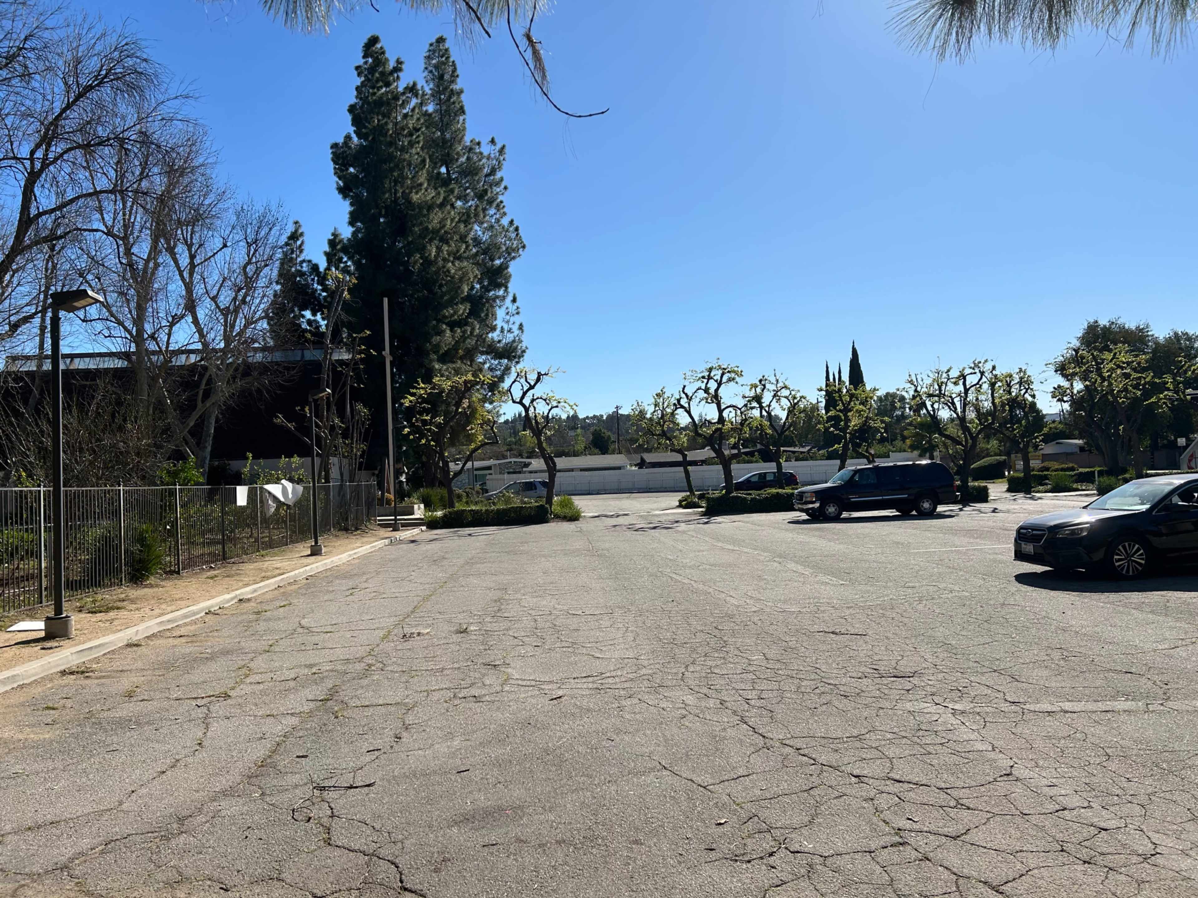 The image shows an empty parking lot with cracked pavement, surrounded by a few trees and a clear blue sky.