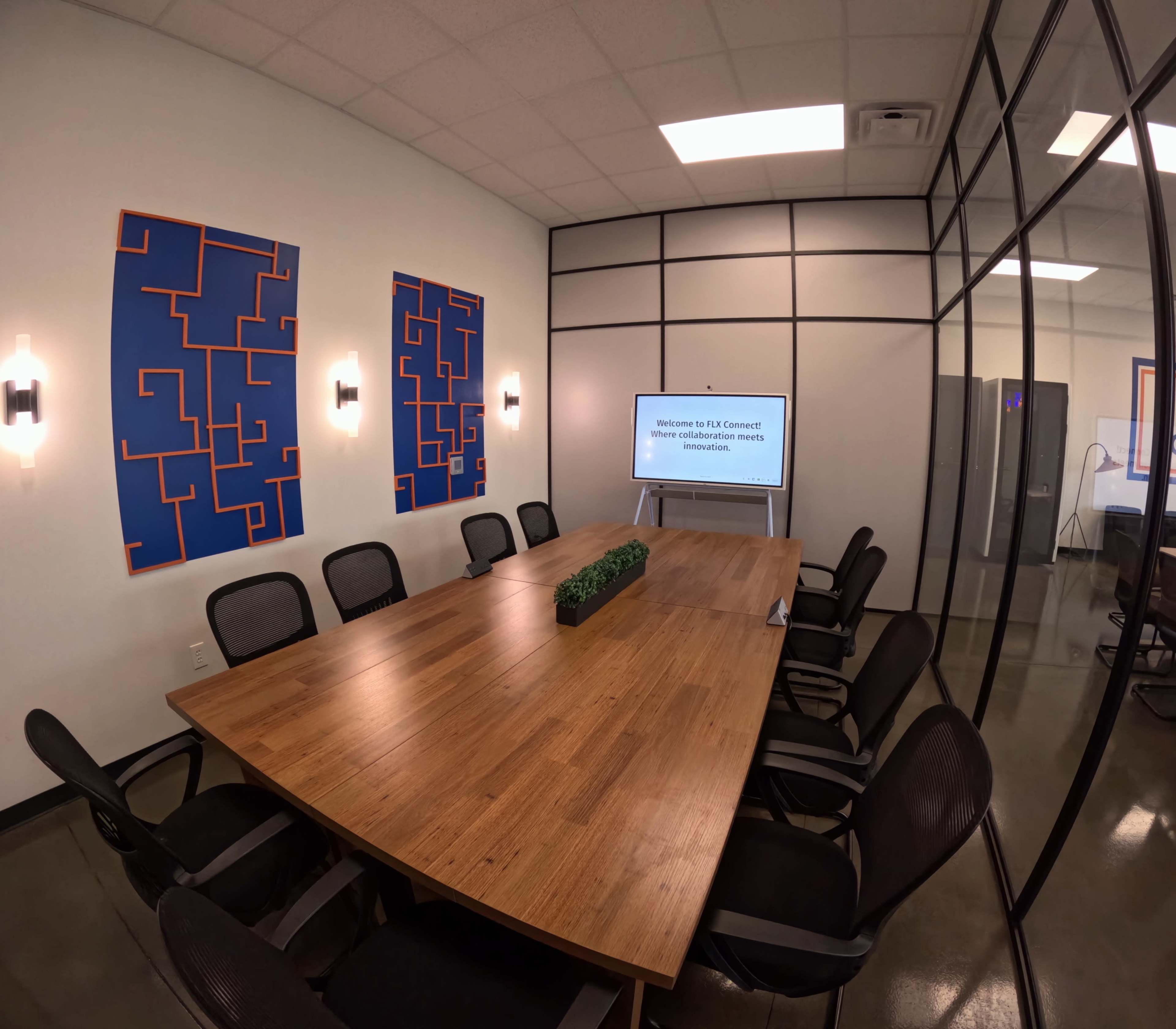 A modern conference room features a large wooden table surrounded by black chairs, with two abstract blue wall art pieces and a presentation screen displaying a welcome message.
