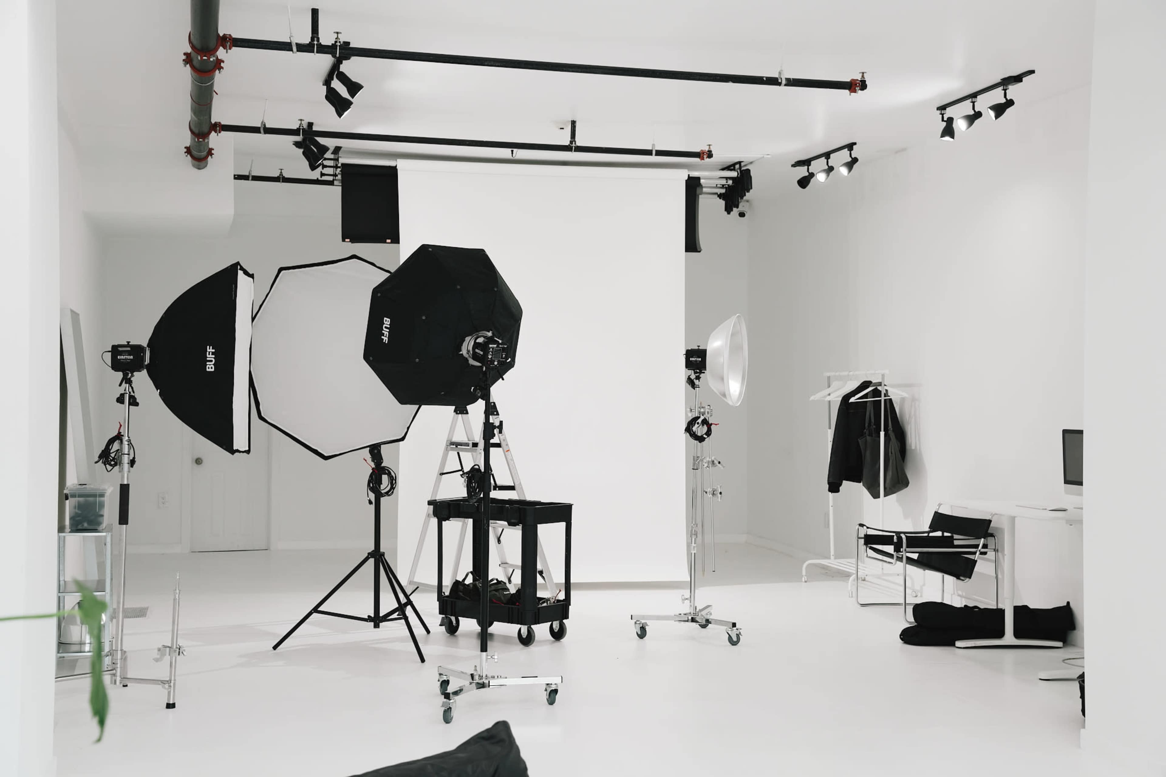 The image shows a bright, minimalistic studio setup with large softbox lights, a seamless backdrop, and various photography equipment arranged in a white room.