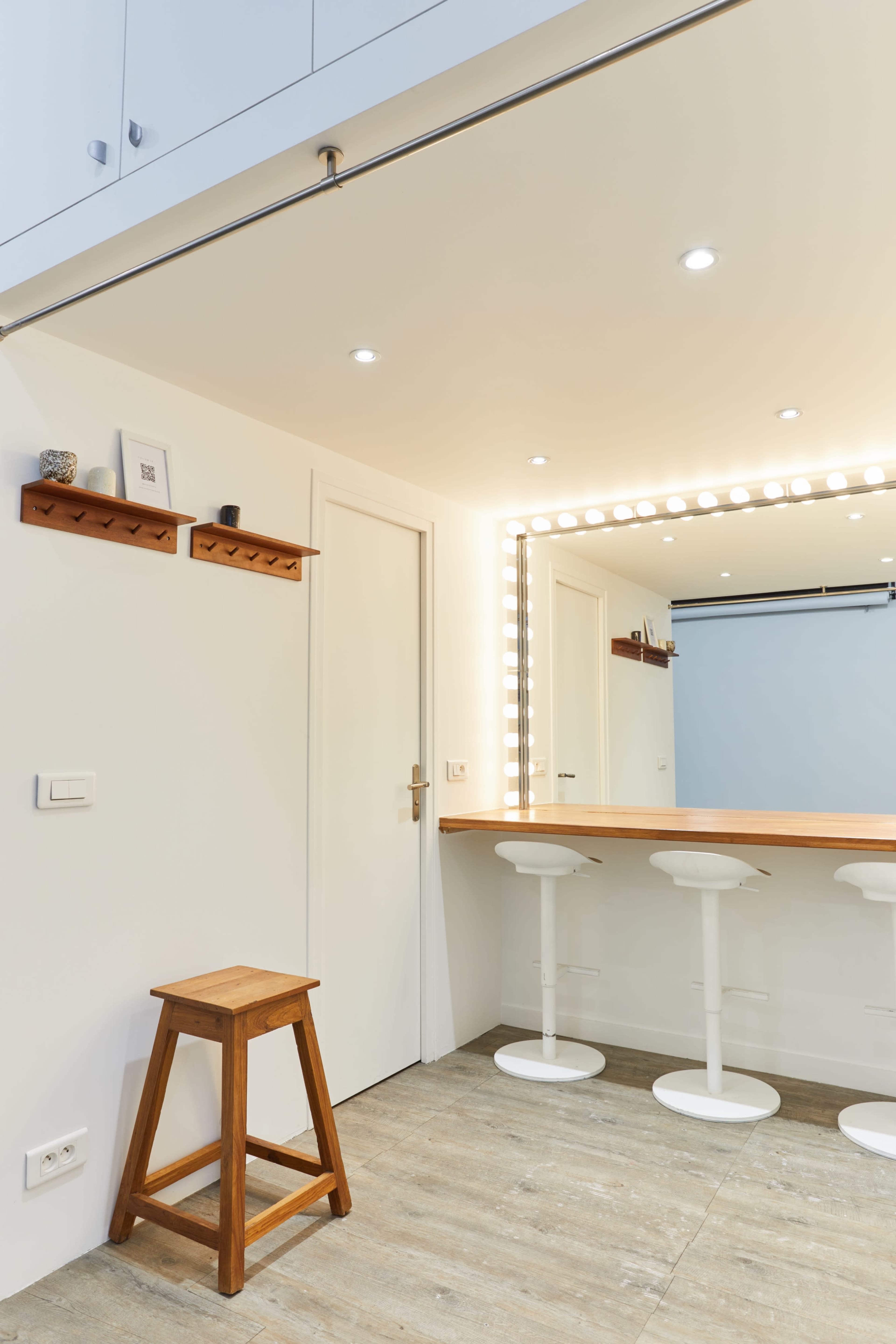 The image shows a modern room featuring a wooden bar with three white stools, illuminated by a lighted mirror and a shelf mounted on the wall.