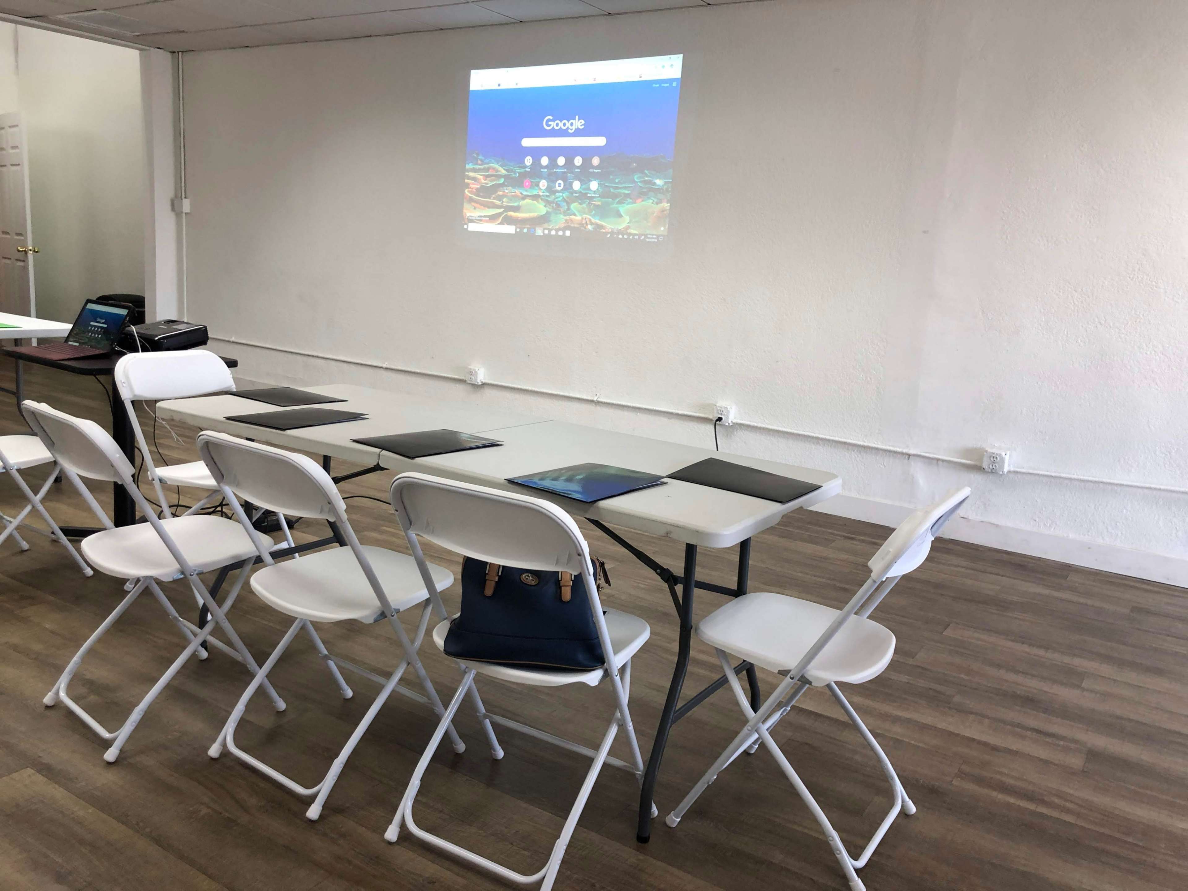 A conference room is set up with a long table and several folding chairs, featuring a projector displaying the Google homepage on a white wall.
