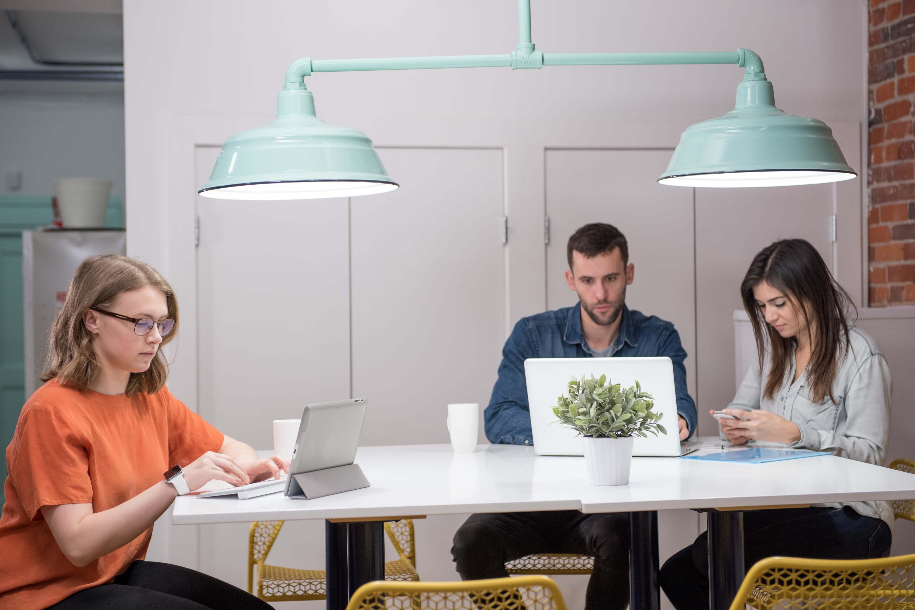 Three people are seated around a table in a modern office space, each using a device: one on a tablet, one on a laptop, and one on a smartphone.