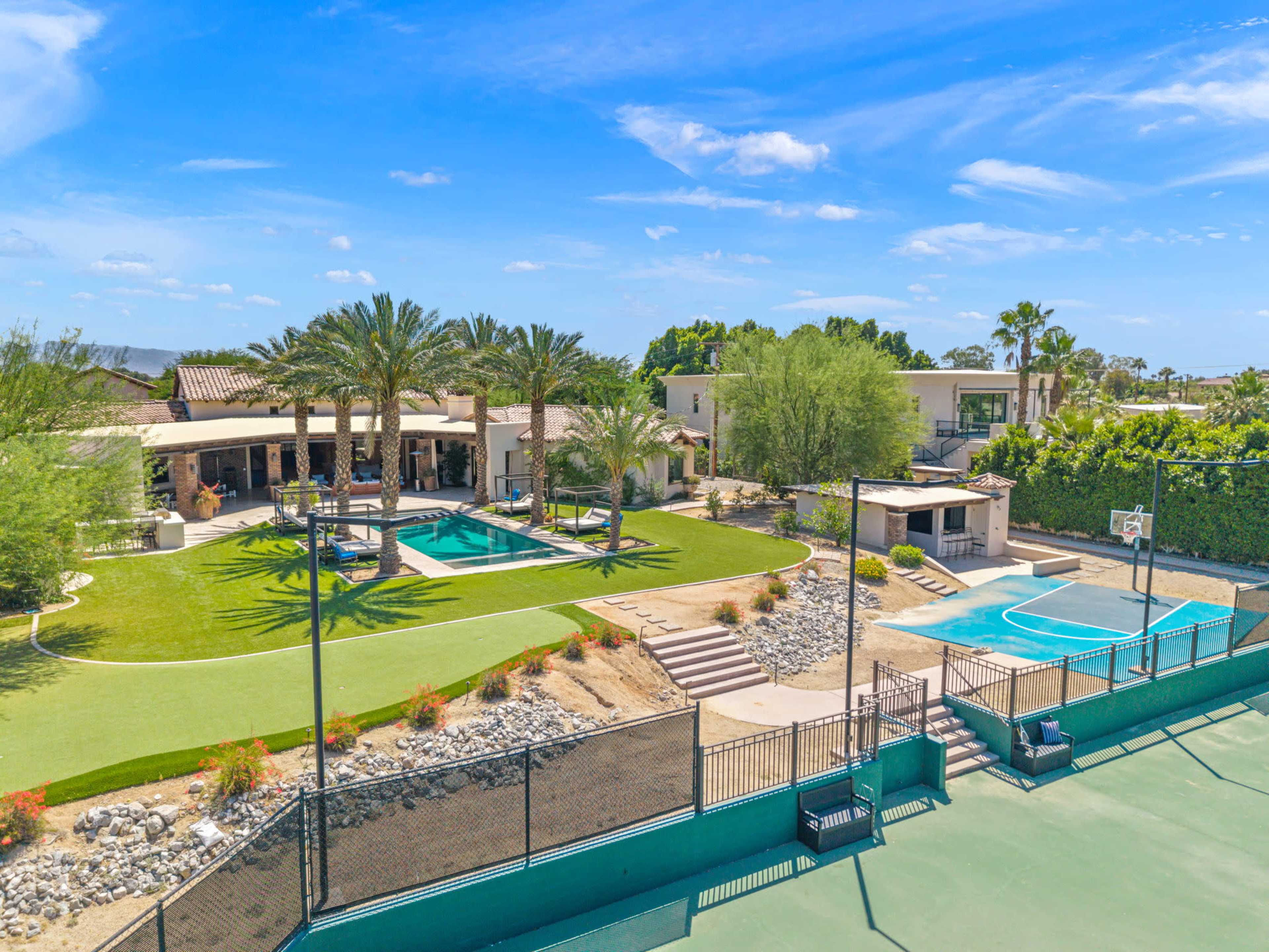 The image shows a spacious residential property featuring a swimming pool, a basketball court, well-maintained lawns, and palm trees under a clear blue sky.