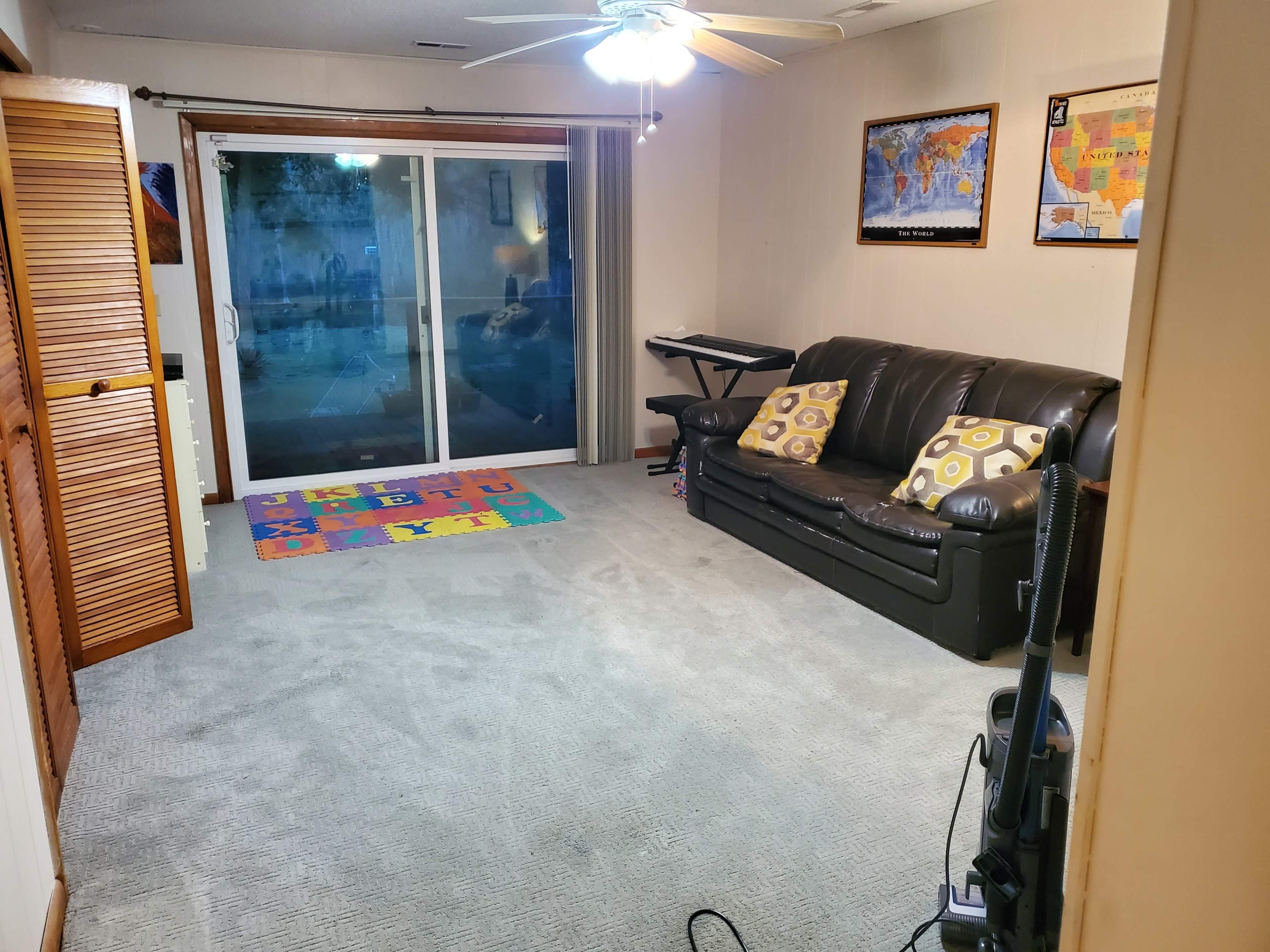 The room features a leather sofa, carpeted floor, a keyboard, a colorful rug, and large sliding glass doors leading to an outdoor area.