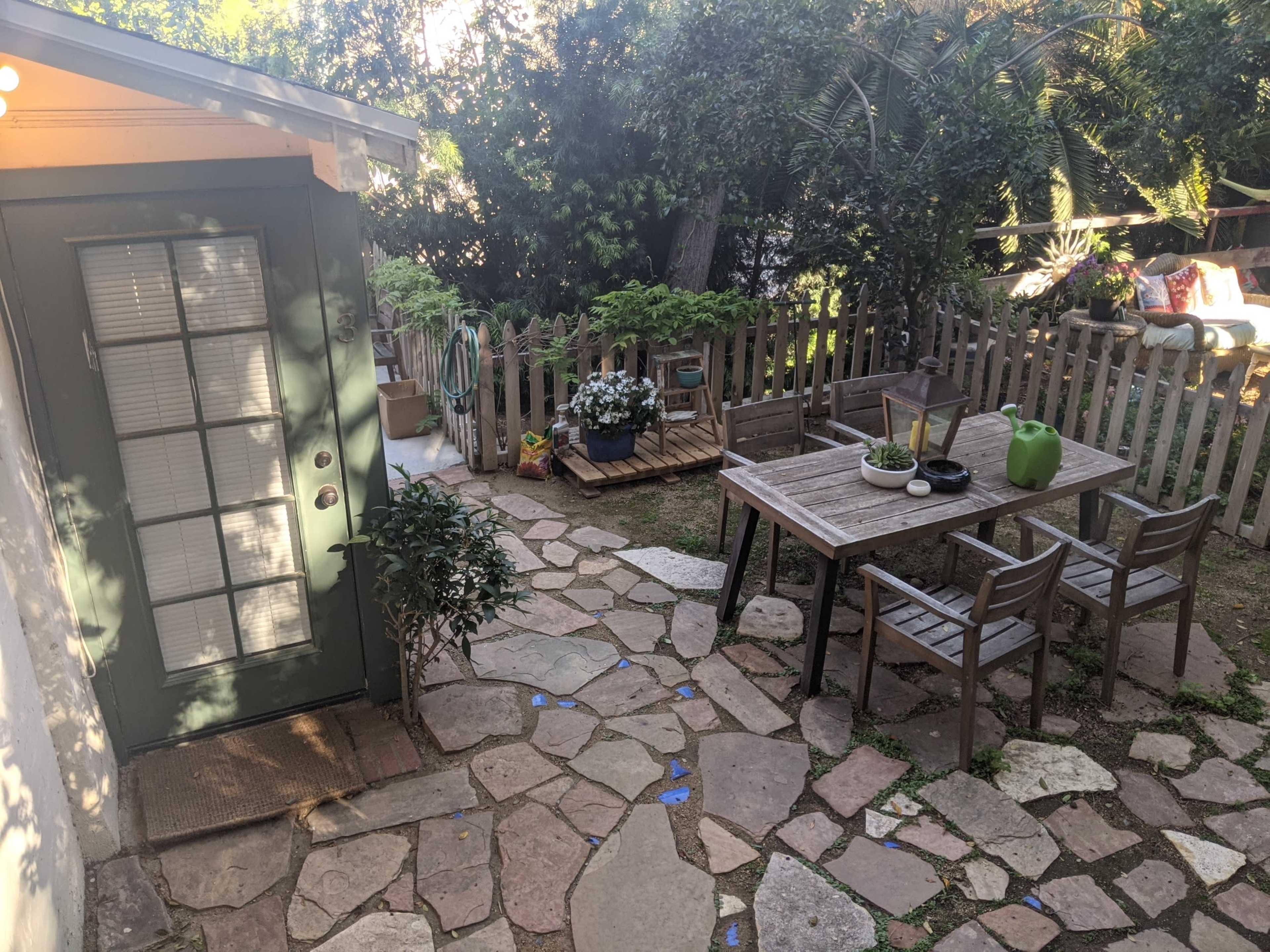 The image shows a backyard with a stone pathway, a wooden table and chairs, a small green building, and various plants surrounded by a wooden fence.