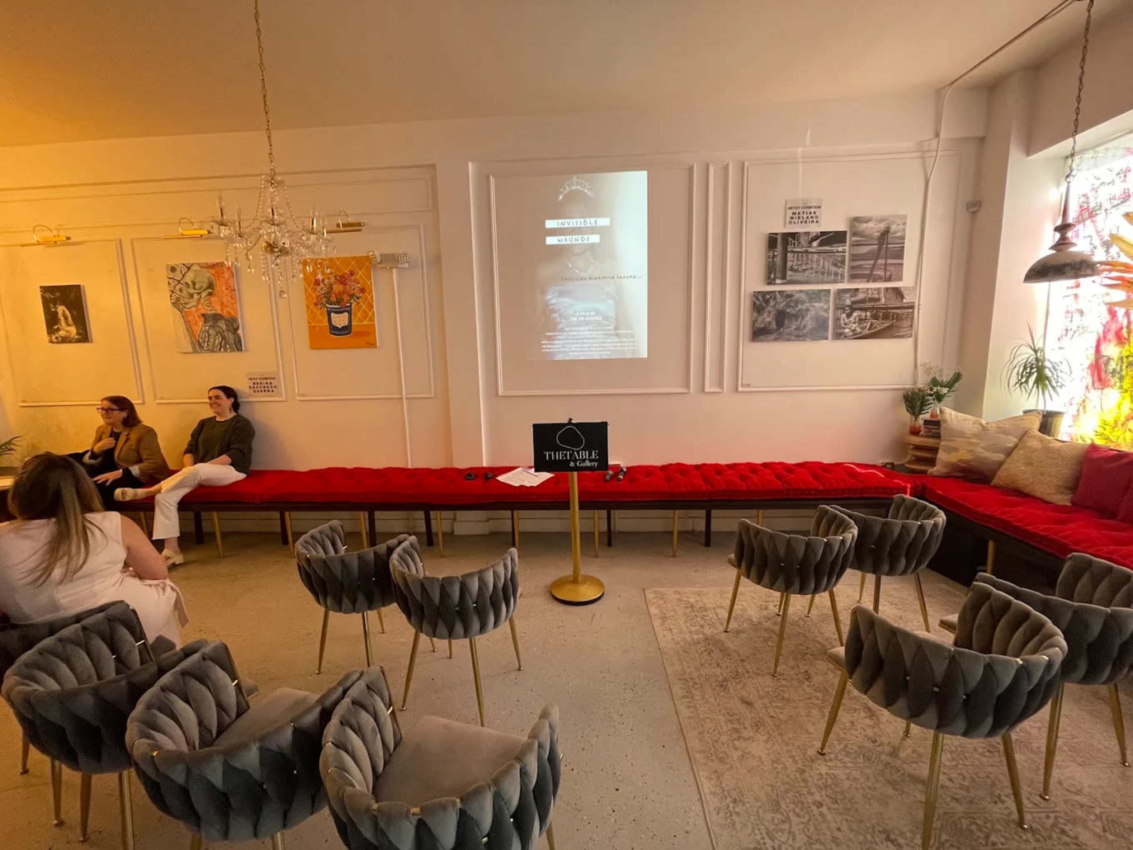 The image shows a modern lounge area with a long red seating arrangement, several velvet chairs arranged in a semi-circle, and walls adorned with framed artwork.