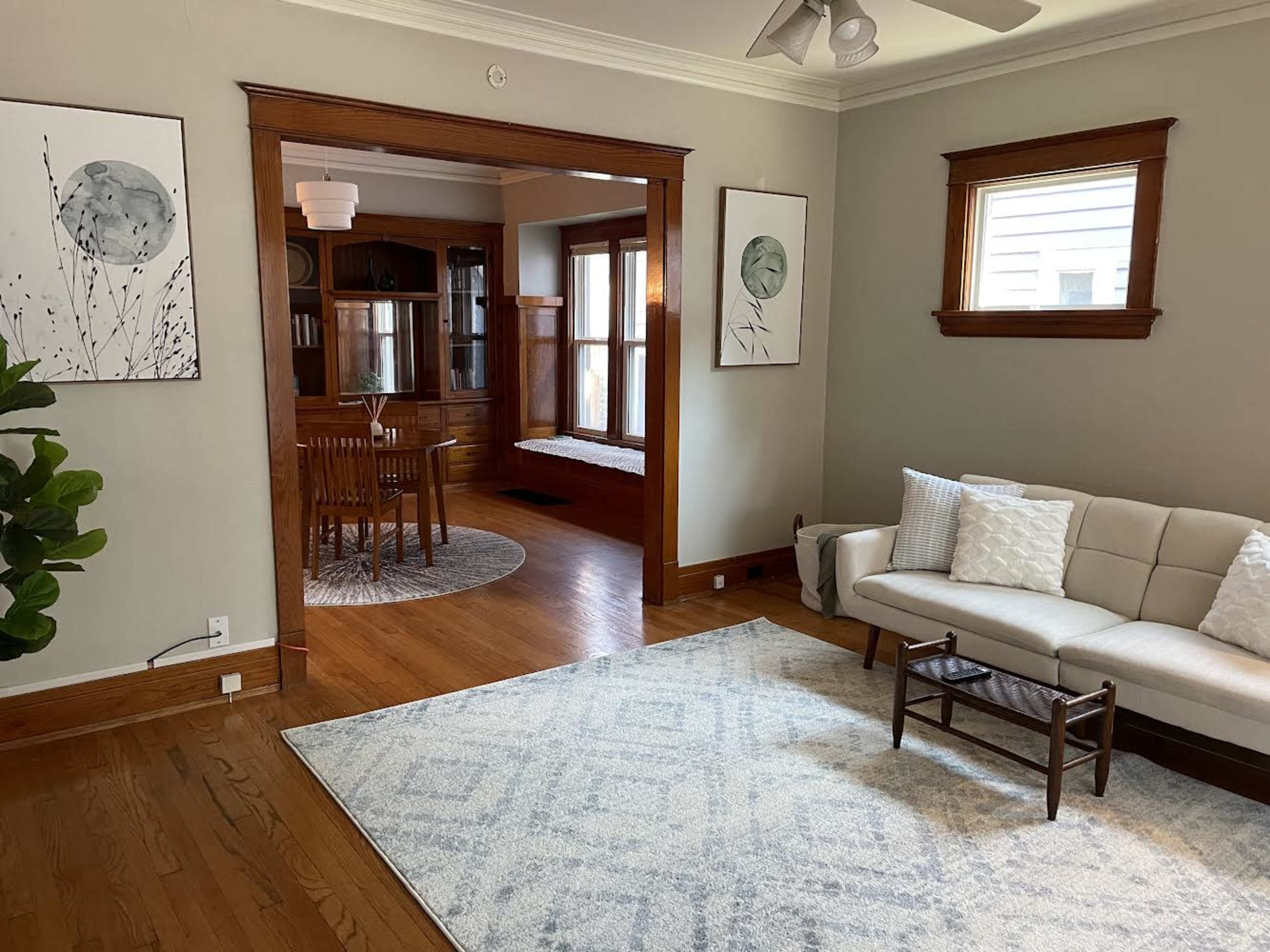 A light-filled living room features a beige sofa, a decorative rug, and two framed botanical prints on the walls, with an adjoining dining area visible through an archway.