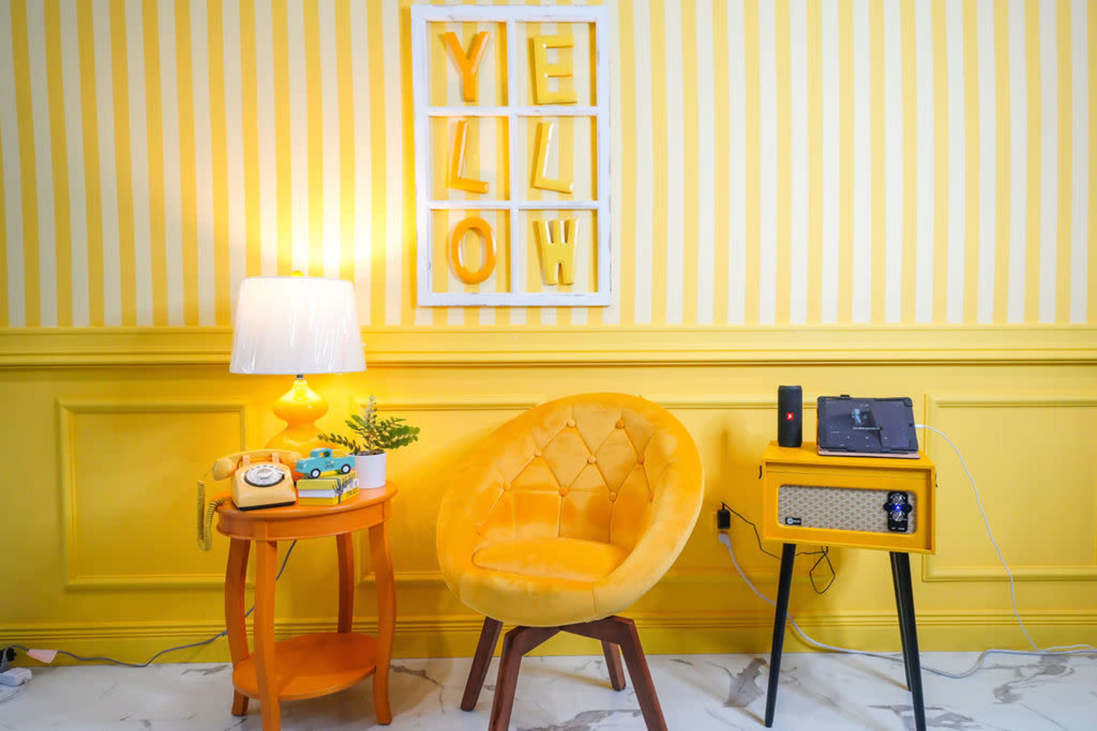 The scene features a brightly decorated room with yellow striped walls, a yellow chair, and decorative elements including a lamp, a plant, and vintage items on the table.