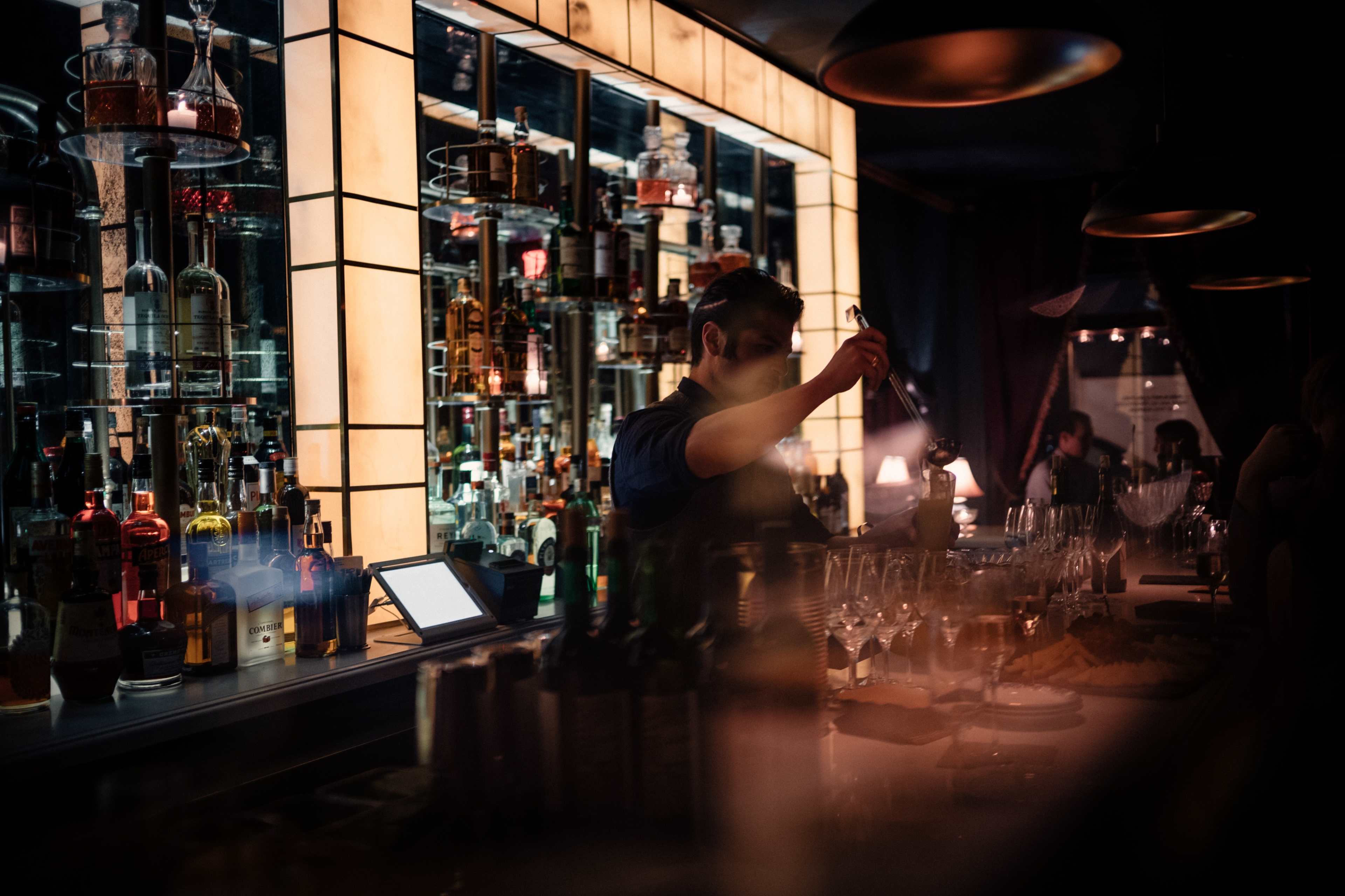 A bartender prepares drinks behind a well-stocked bar illuminated by backlit shelves filled with bottles.