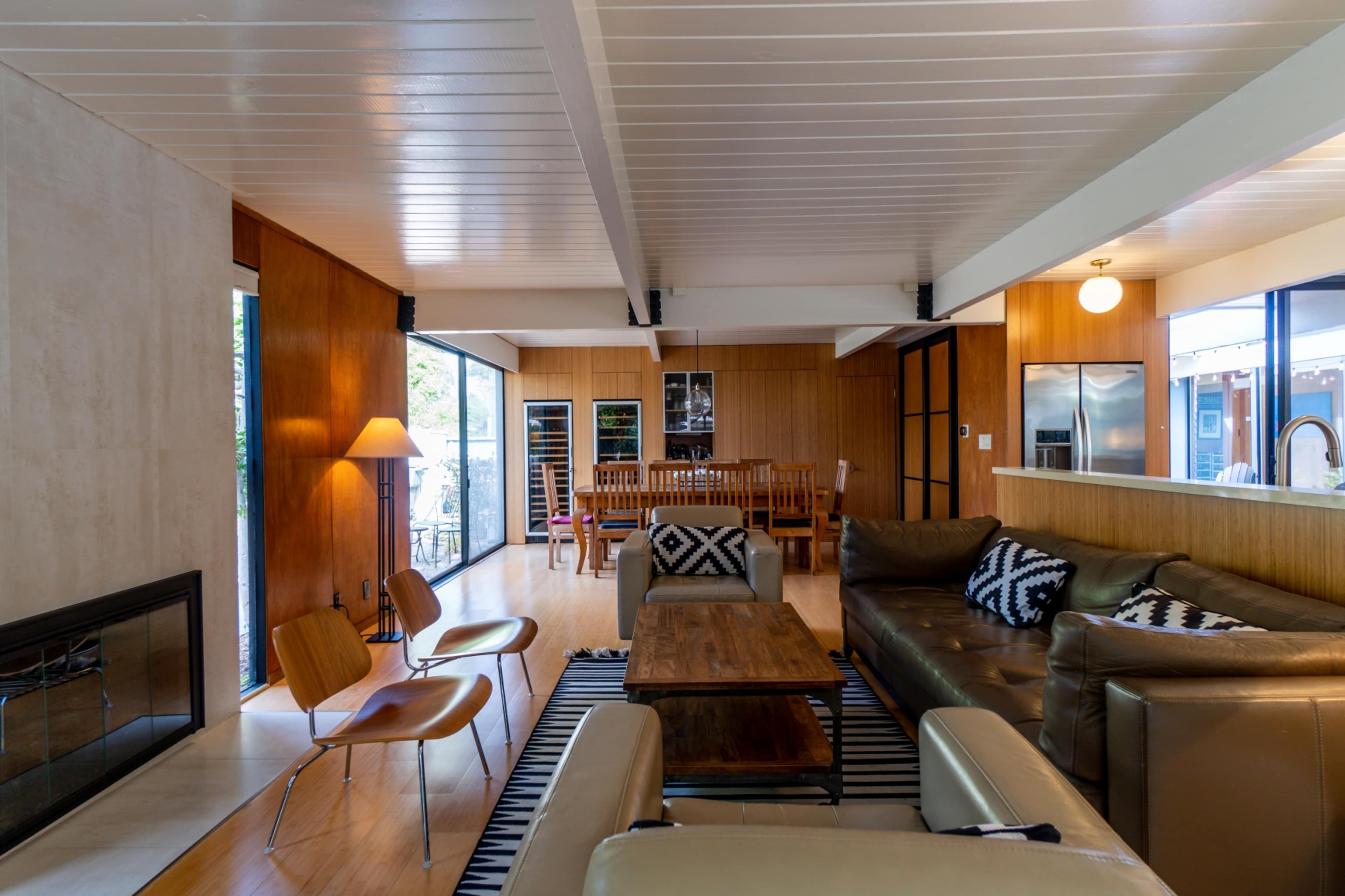 The image shows a modern interior living space with a brown leather sectional sofa, a wooden coffee table, a dining area in the background, and large windows overlooking an outdoor patio.