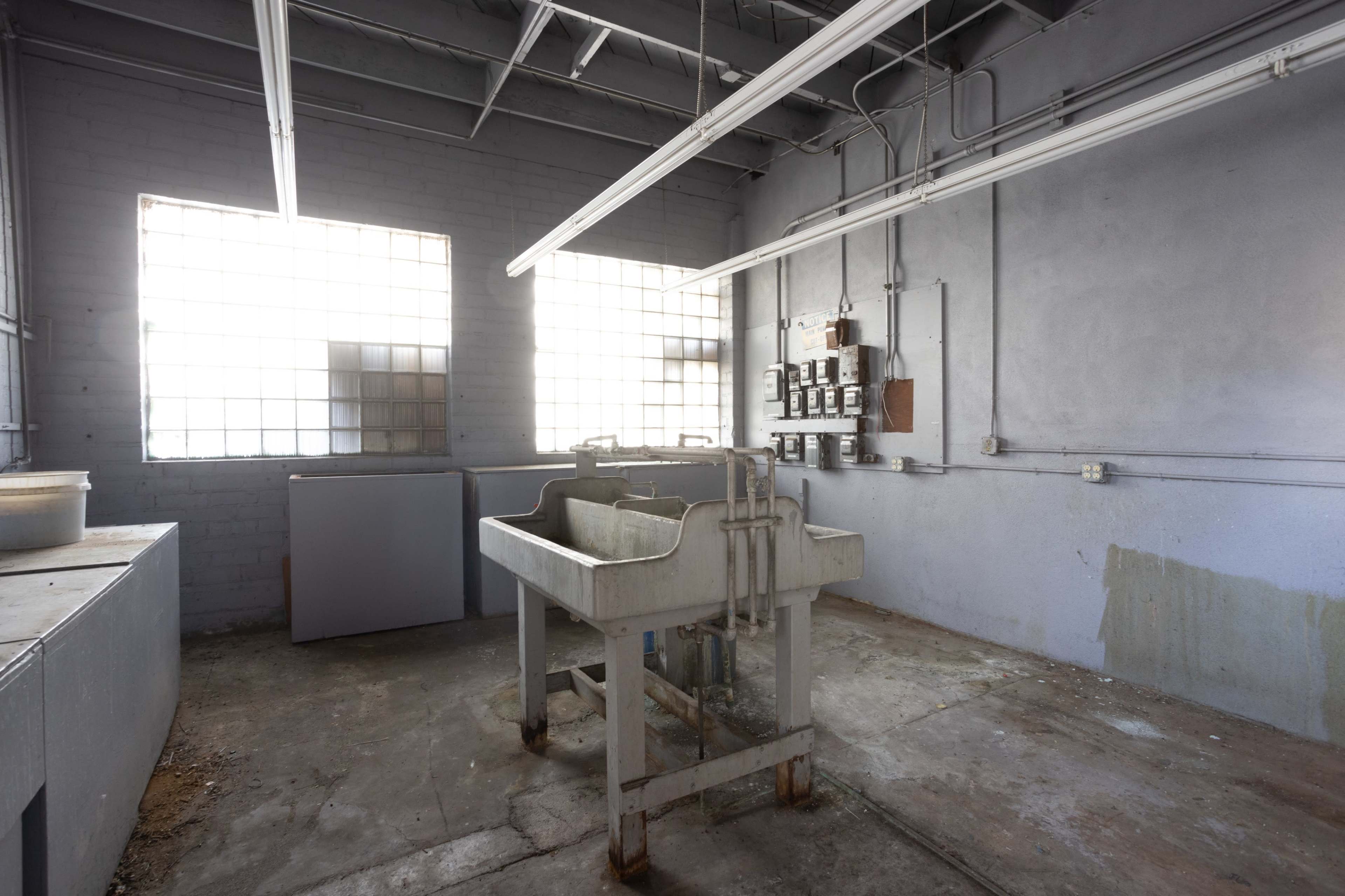 A vacant industrial room with a large, white sink in the center, surrounded by gray walls and windows with grid patterns.