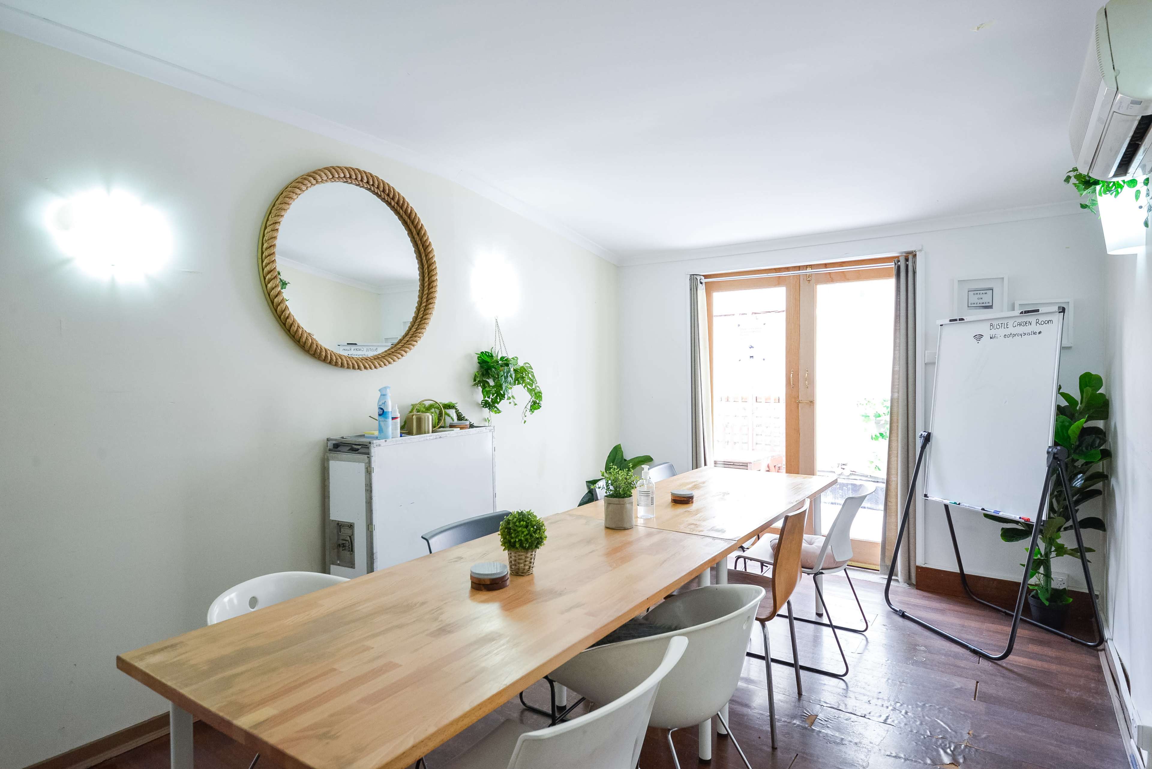 A bright meeting room features a wooden table surrounded by chairs, a mirror on the wall, and a whiteboard near a window.