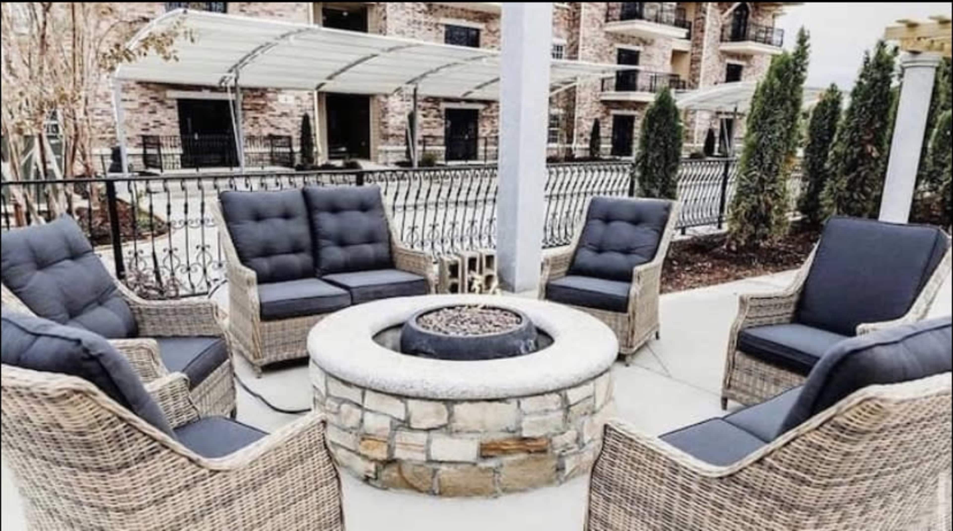A circular stone fire pit surrounded by six wicker chairs with dark cushions, arranged on a patio.