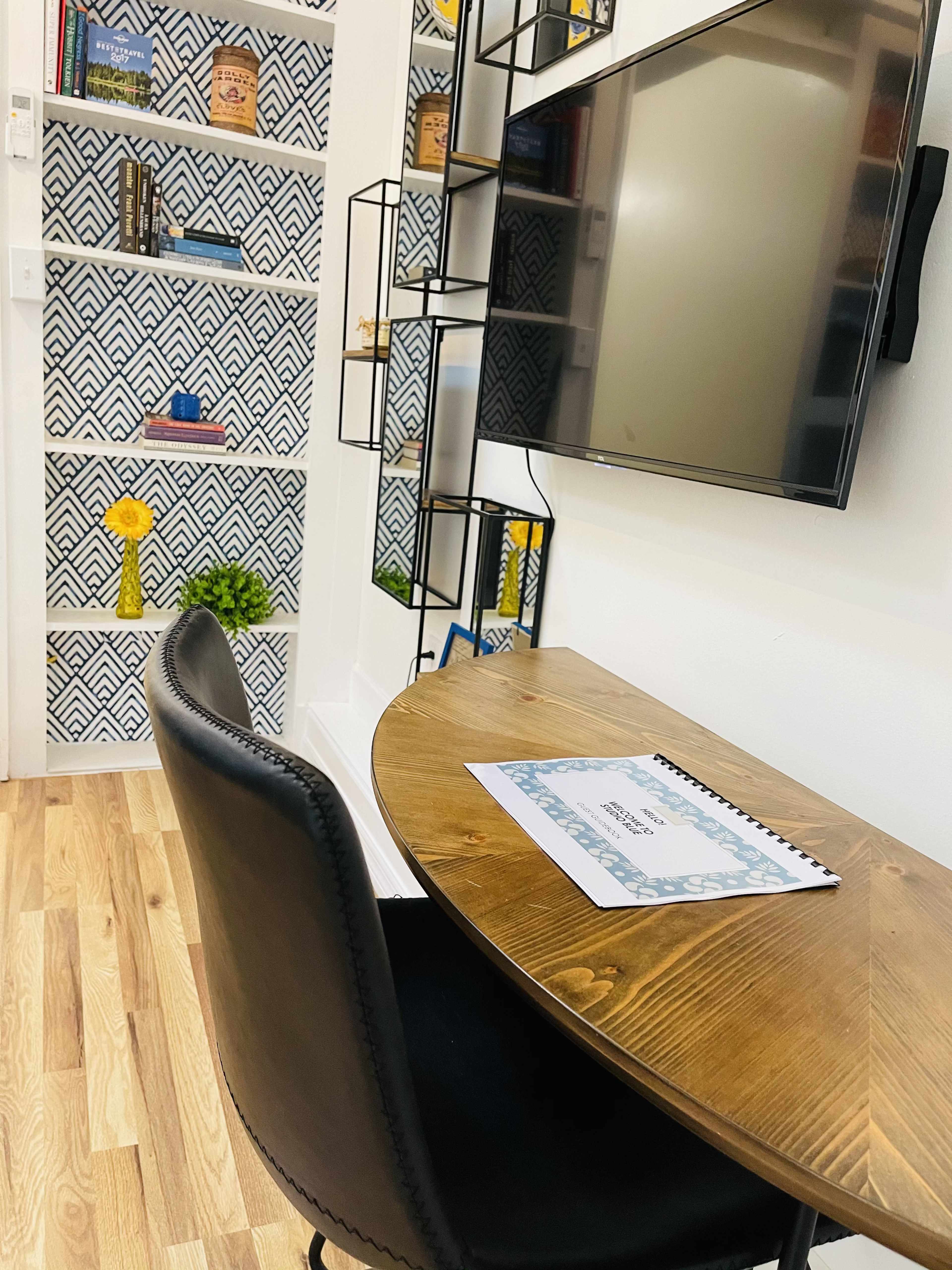 A small workspace with a round wooden table, a black chair, and a wall-mounted TV, featuring patterned shelving in the background.