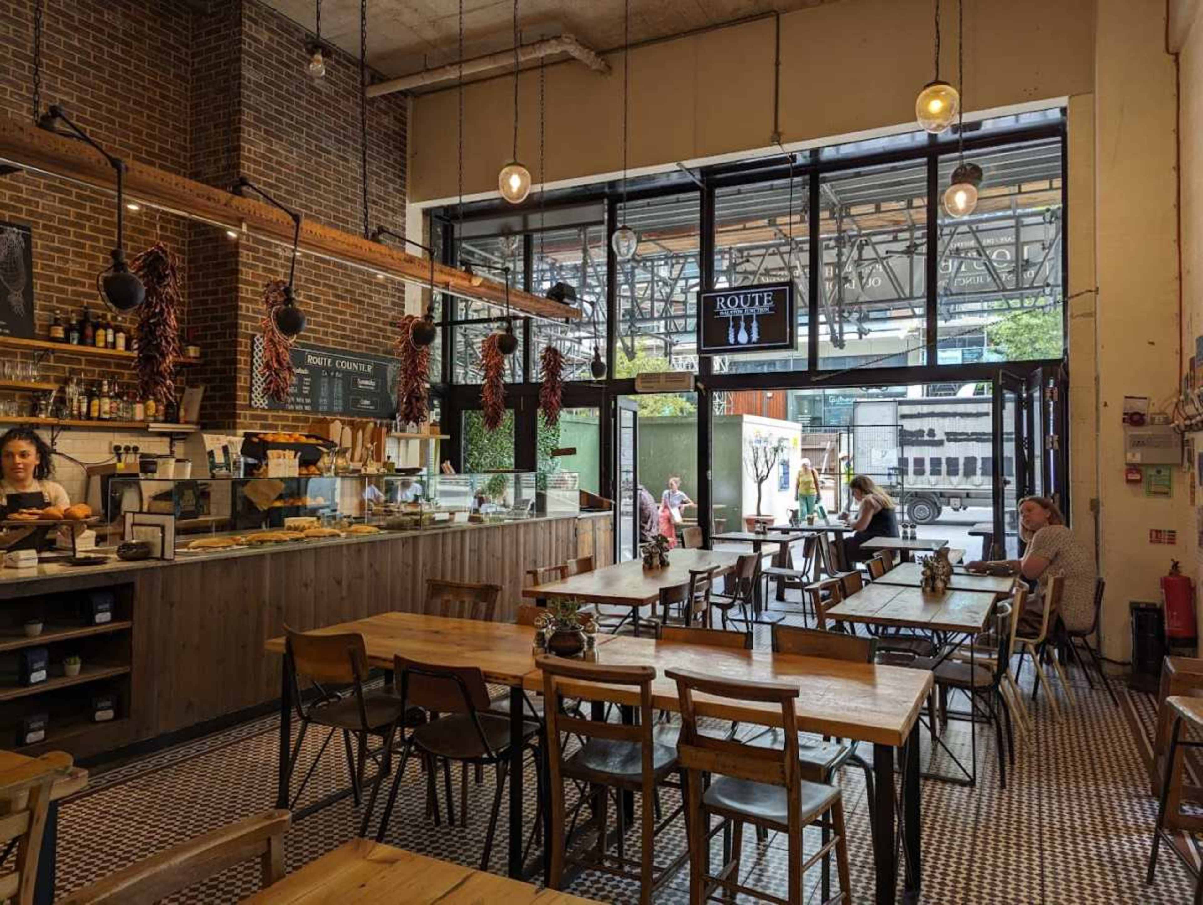 The image shows a modern cafe interior with wooden tables, a counter displaying food, and large windows providing natural light.