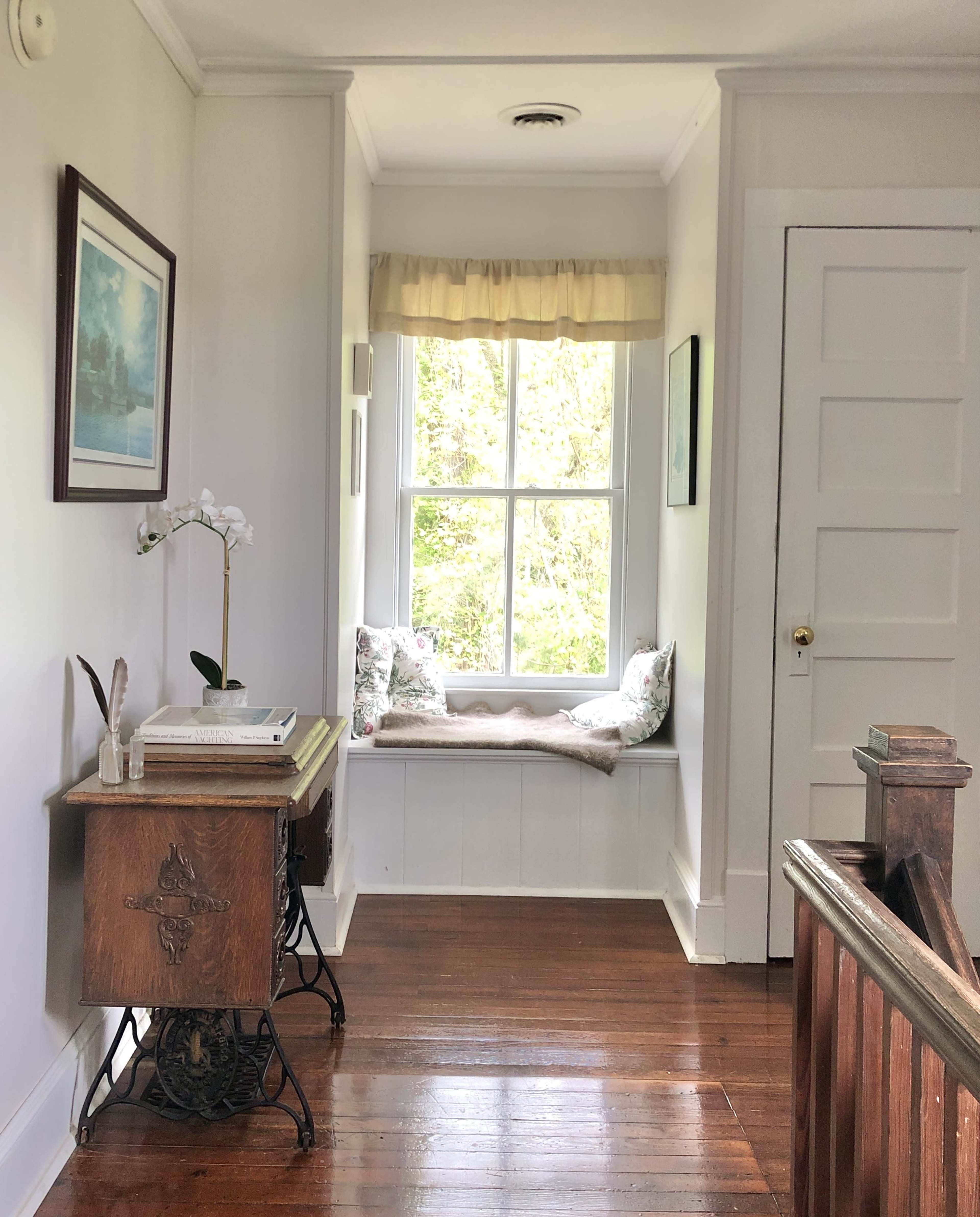 A hallway features a wooden window seat with decorative cushions, a small table, and a door leading to another room.