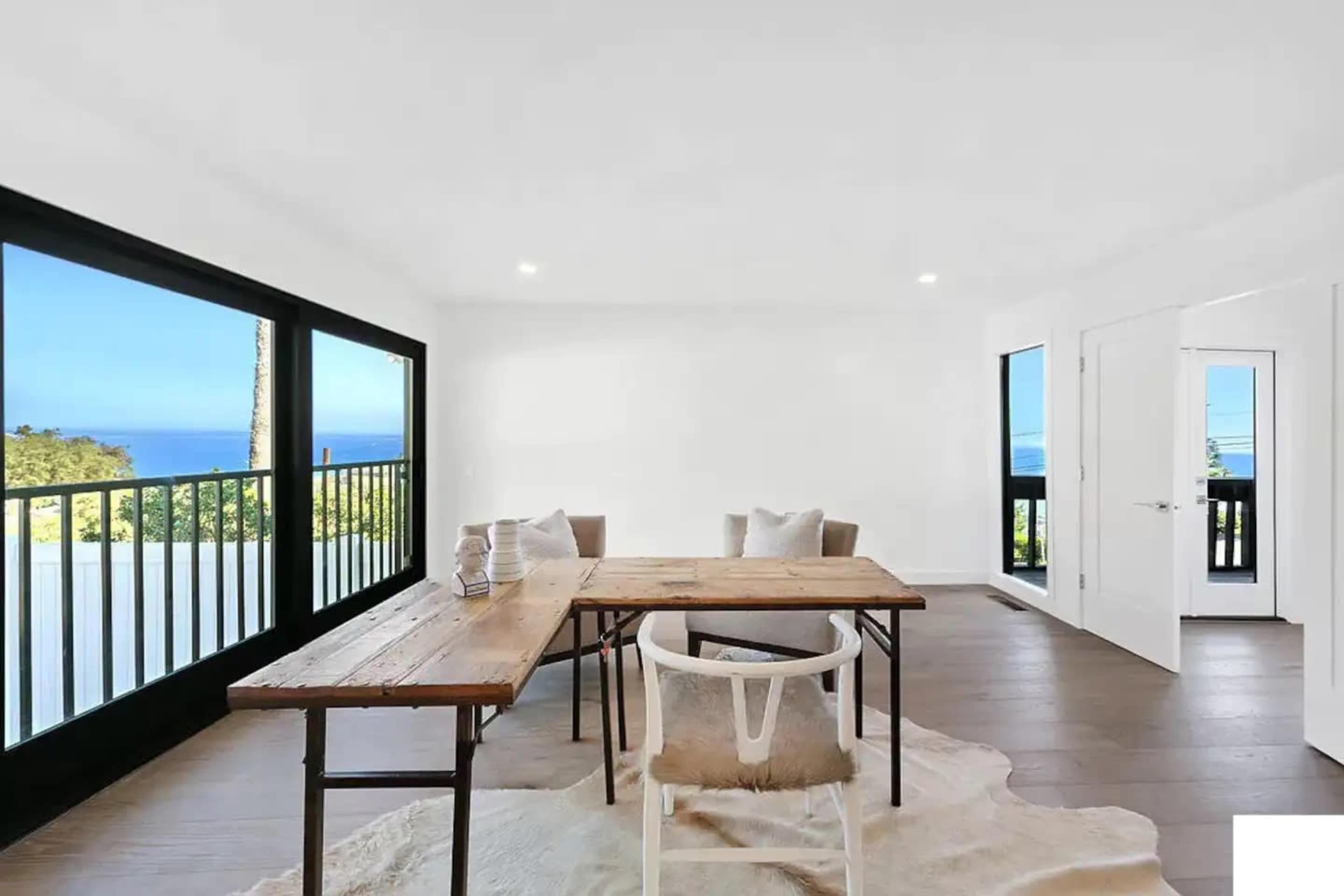 A minimalist dining area features a wooden table surrounded by chairs, with large windows offering a view of the ocean.
