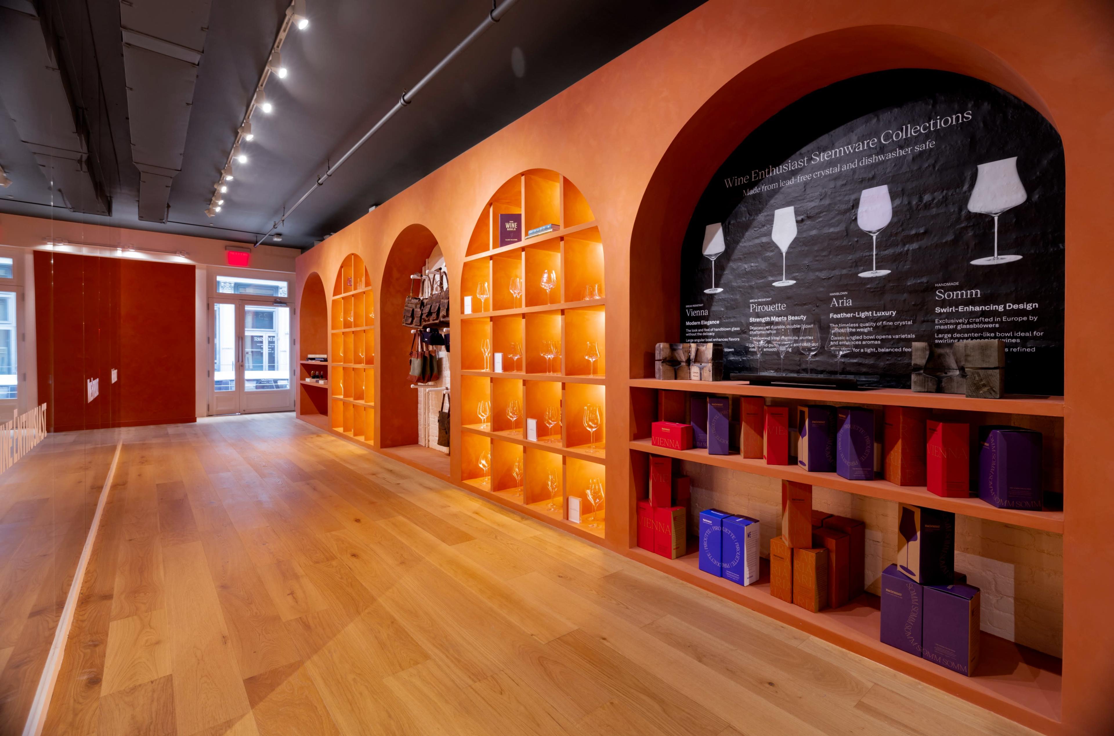 The image shows a modern retail space with orange walls, arched shelves filled with stemware collections, and a large black wall graphic.
