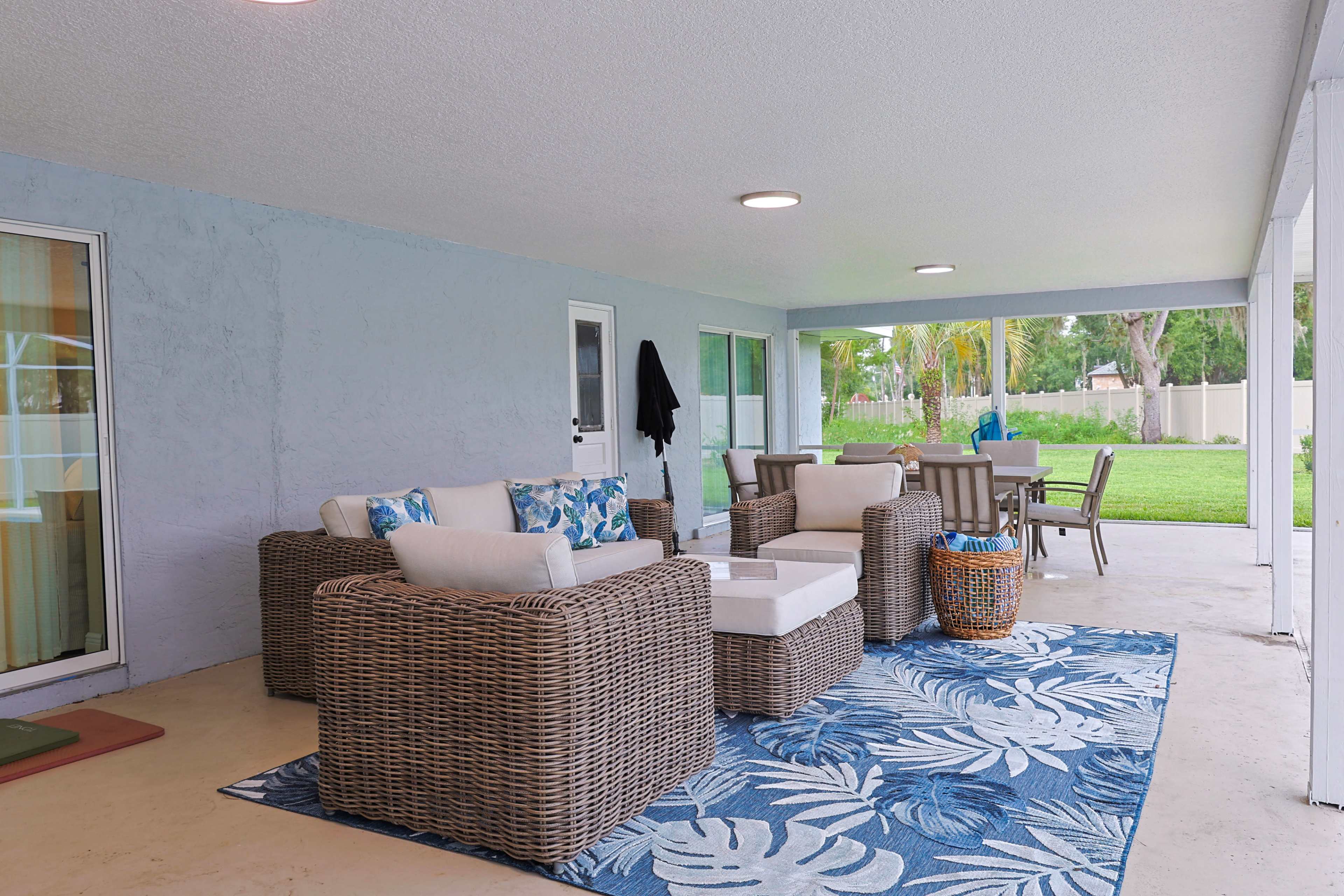The image shows a covered outdoor patio area featuring a seating arrangement with wicker furniture, a coffee table, and a patterned rug, overlooking a green lawn.