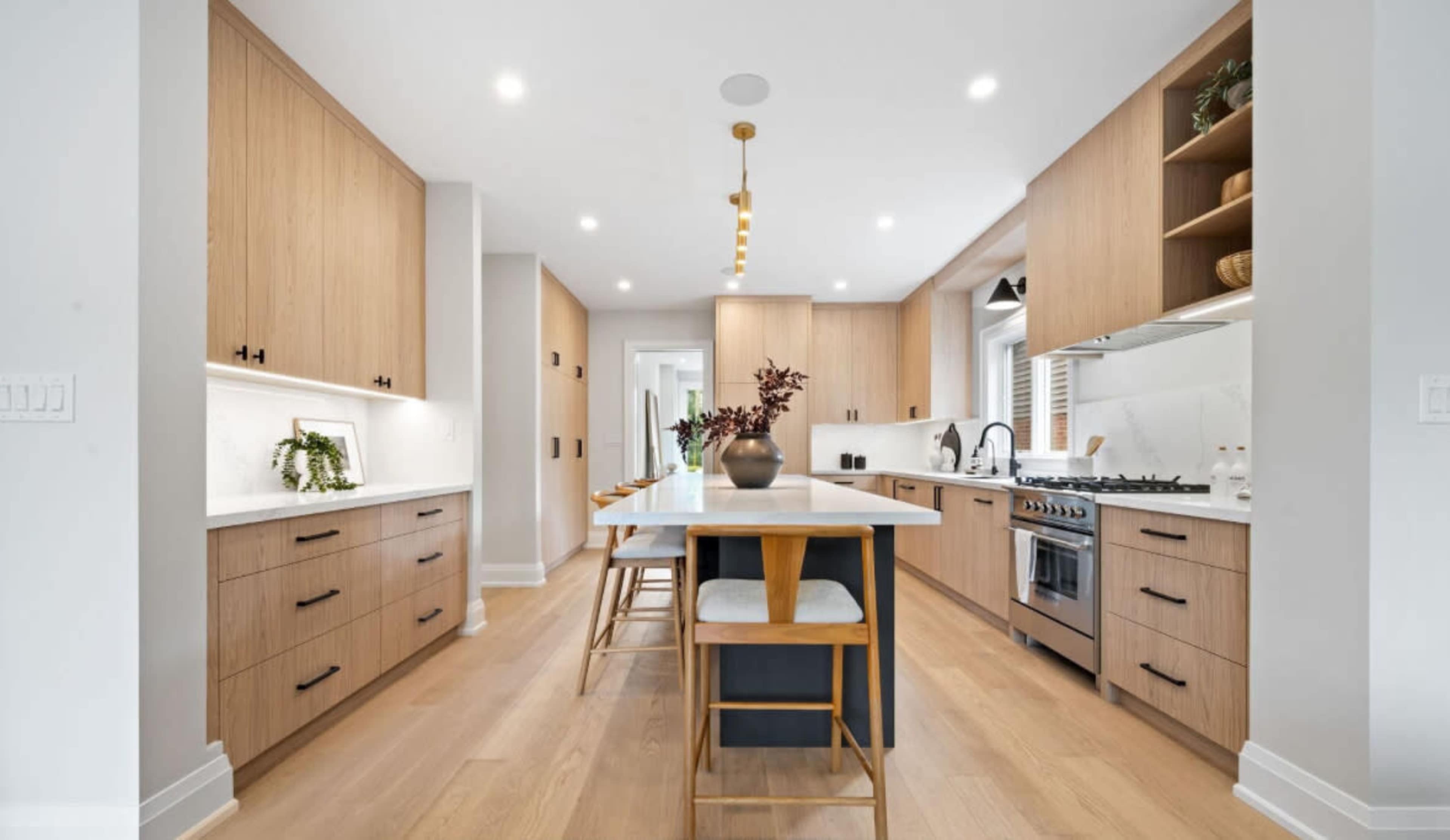 The image shows a modern kitchen with light wood cabinets, a large island with seating, and stainless steel appliances.