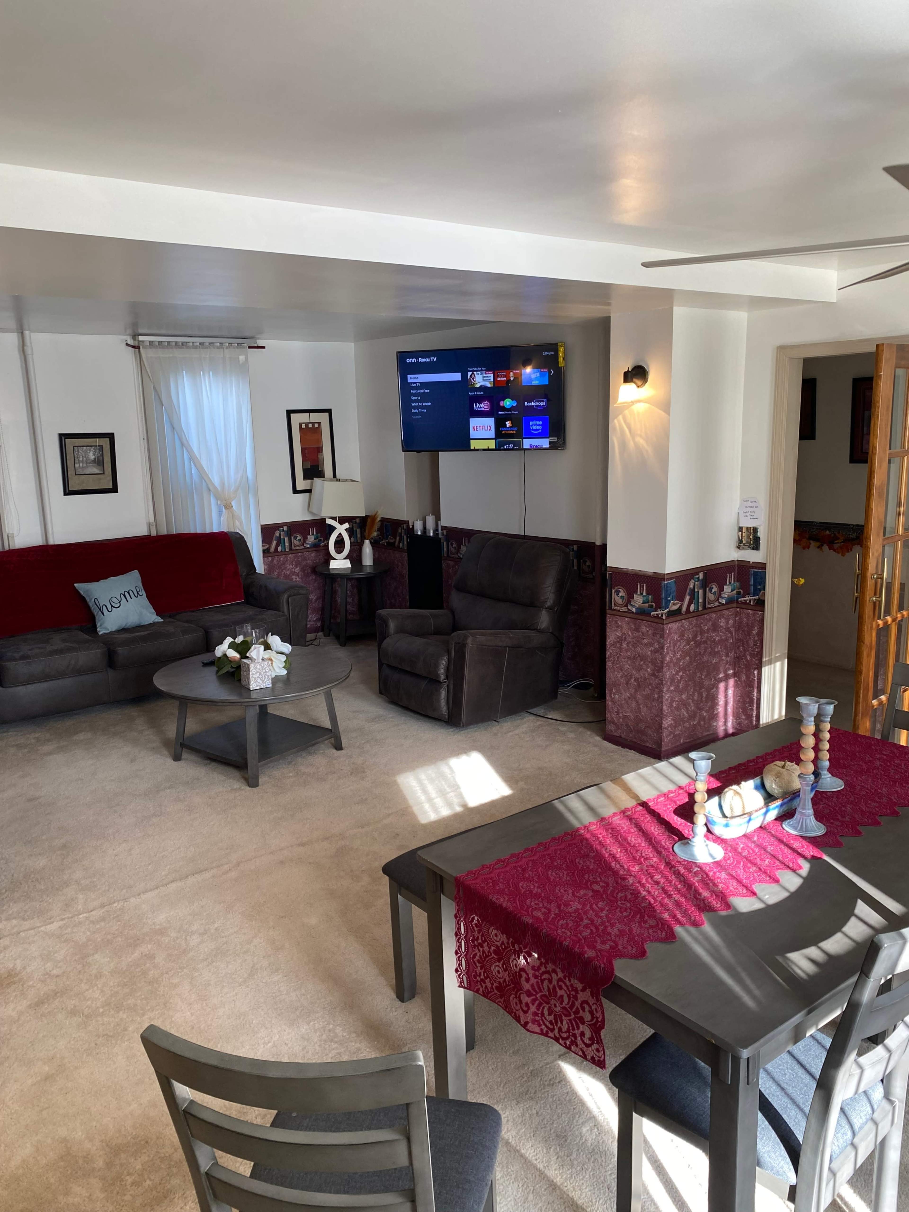 The image shows a cozy living room with a seating area, a television mounted on the wall, and a dining table with decorative candles.