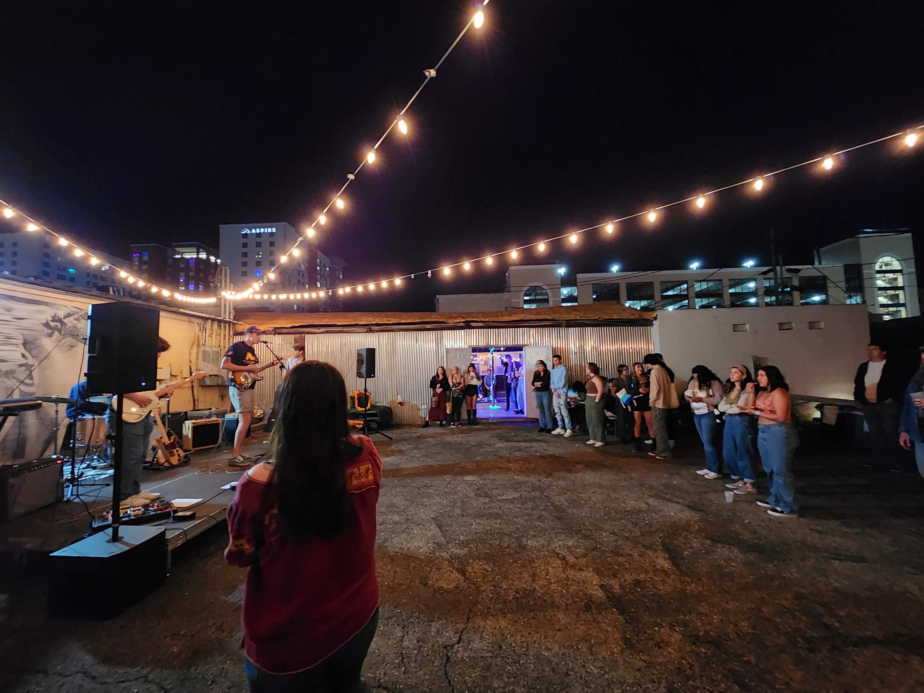 A musician performs on a rooftop stage in front of a small crowd under string lights at night.