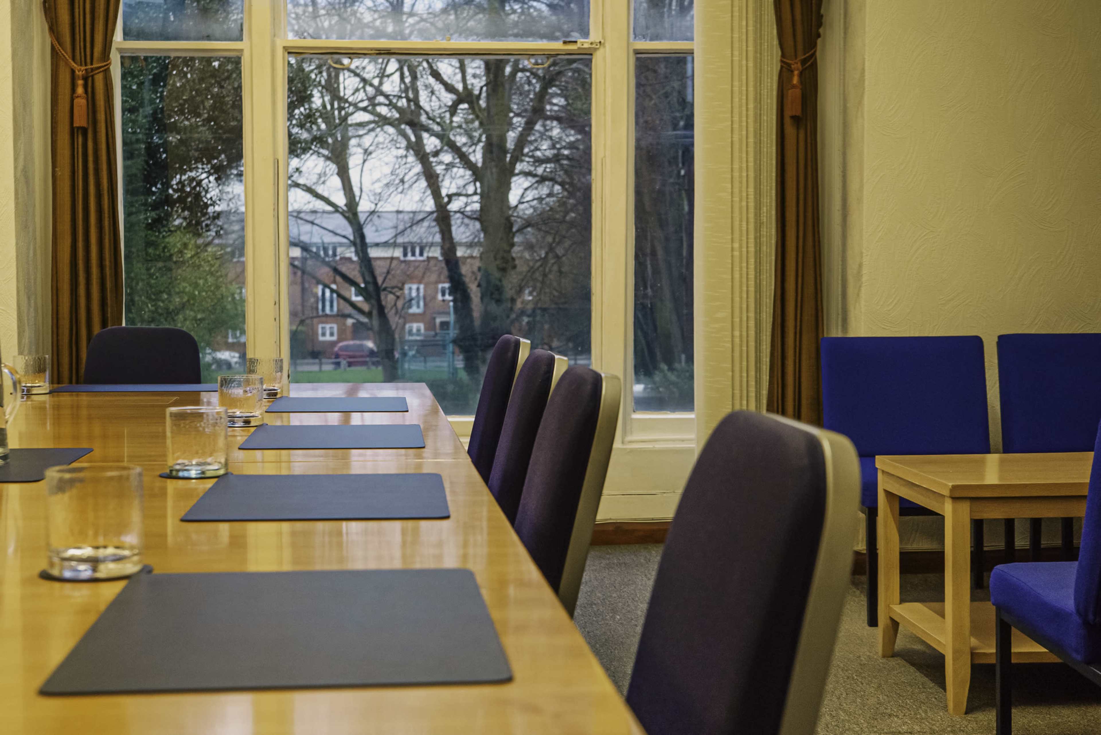 A conference room with a long wooden table set with placemats, chairs on one side, and large windows revealing a view of trees and buildings outside.