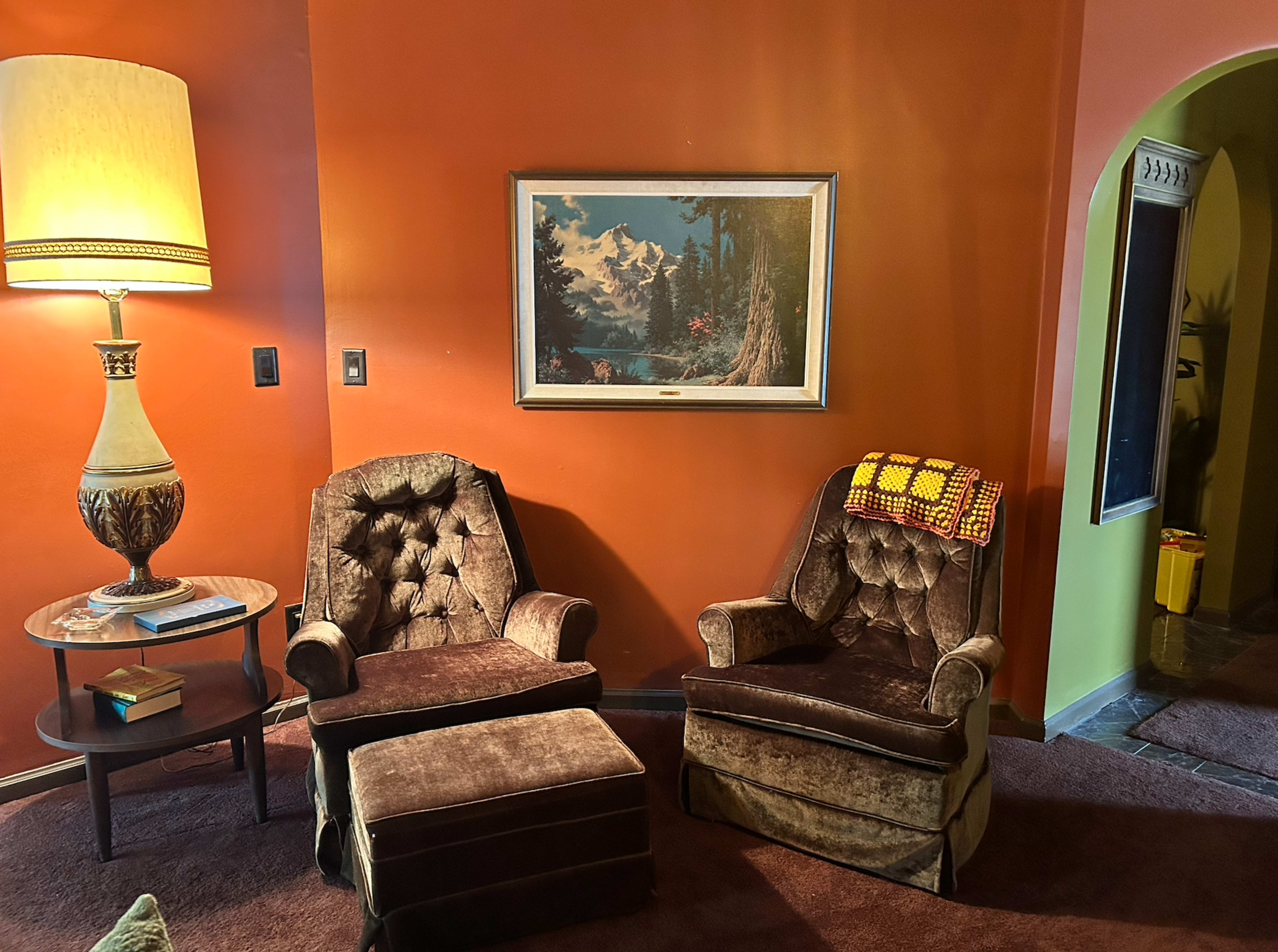 The image shows a cozy living room with two upholstered chairs, an ottoman, a lamp, and a landscape painting on an orange wall.