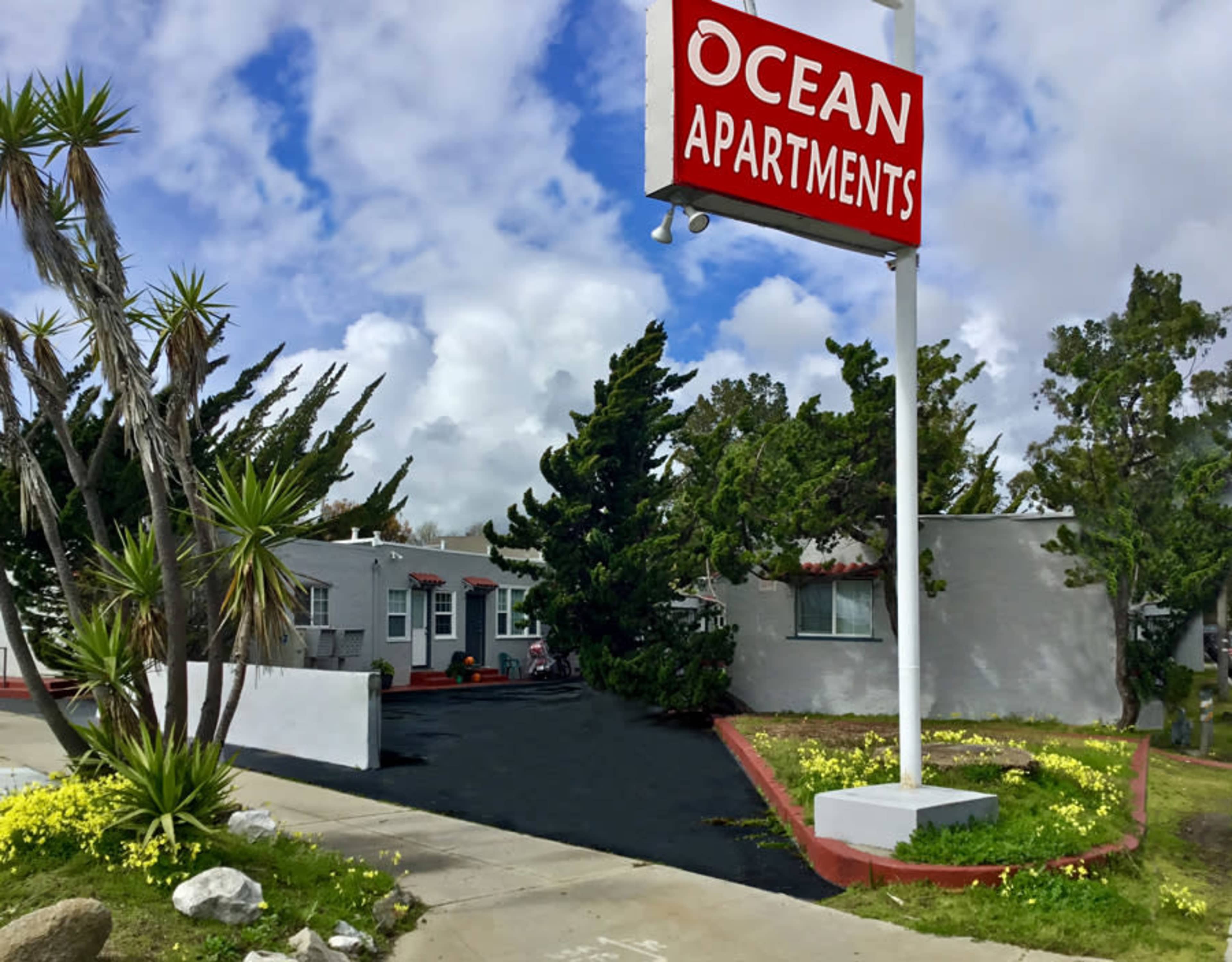A sign reads "Ocean Apartments" outside a small complex with several white buildings surrounded by greenery and a paved walkway.