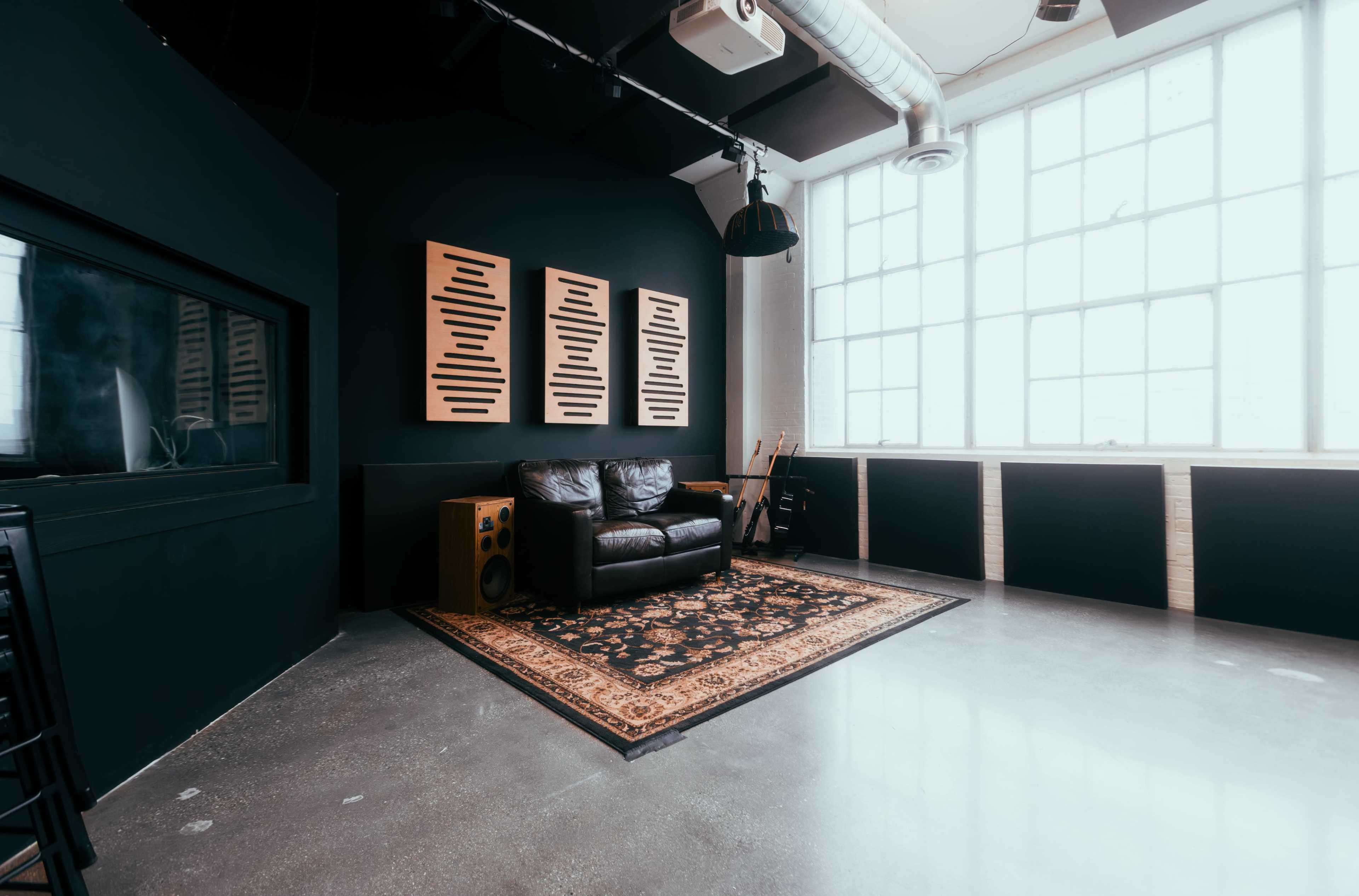 The image shows a modern studio space featuring a black couch on an ornate rug, with large windows and soundproof panels on the walls.