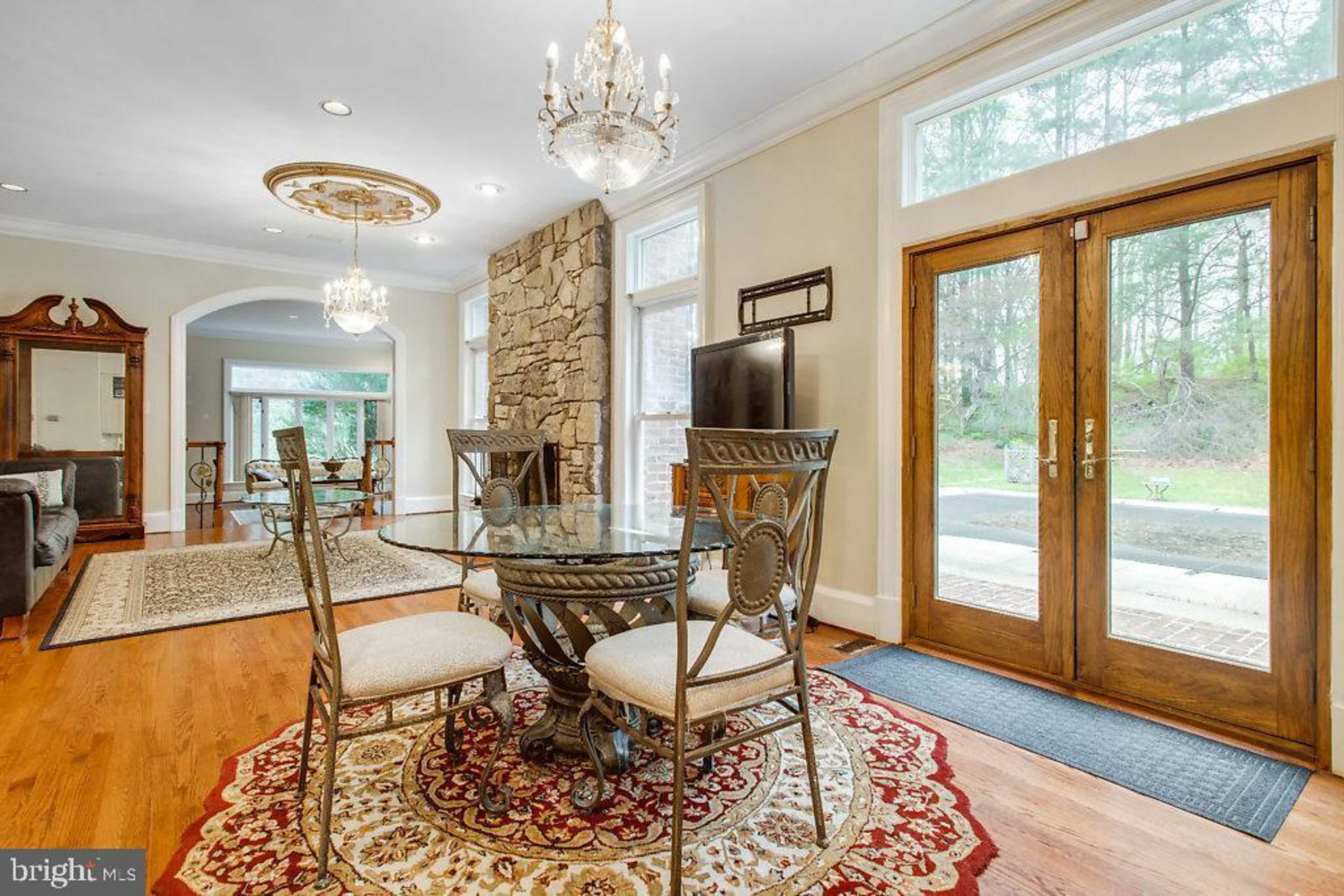 A dining area with a round glass table and four chairs on a patterned rug, illuminated by a chandelier and featuring large windows and double front doors.