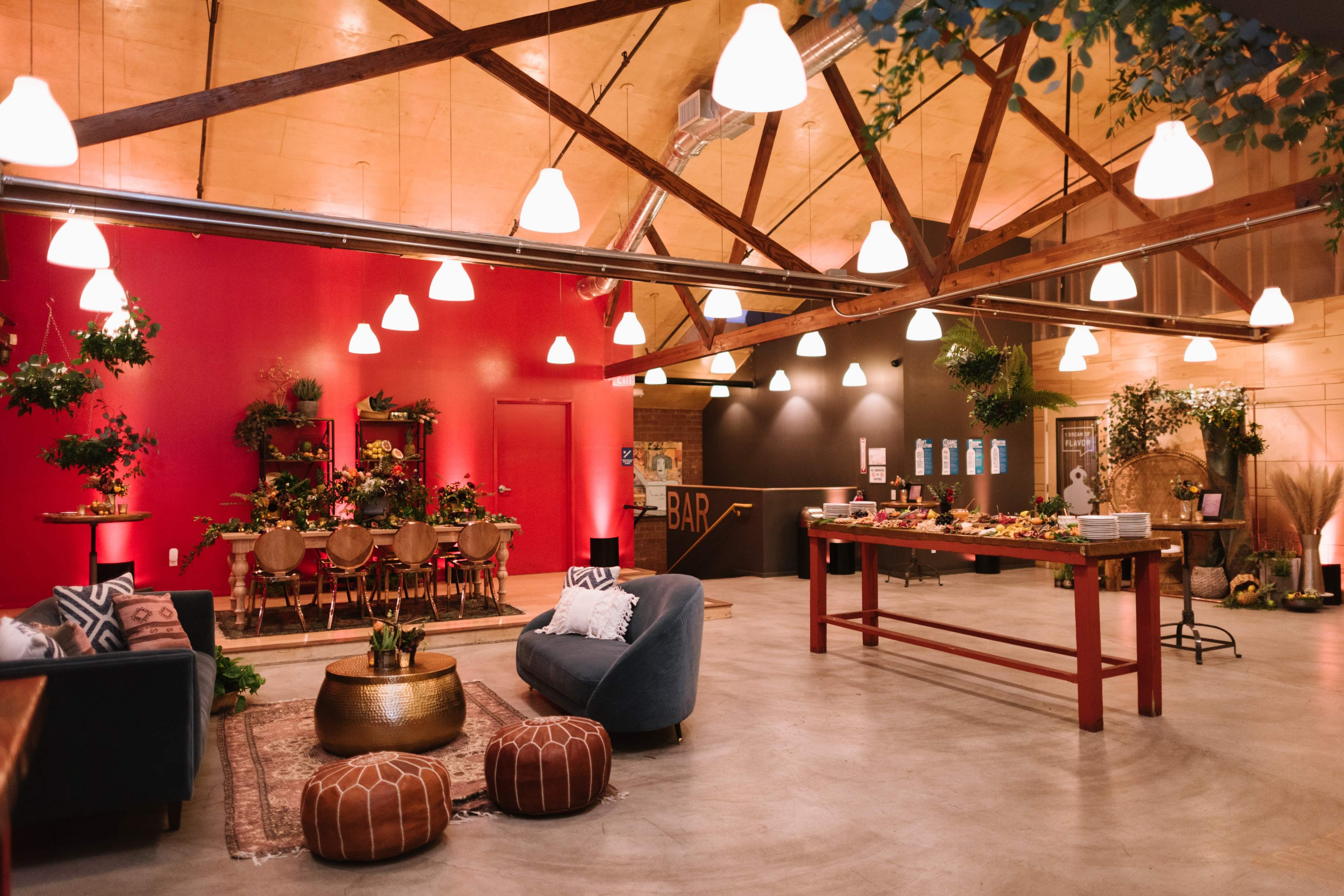 An indoor event space featuring a bar area with seating, a central table adorned with decorations, and warm lighting from hanging lamps.