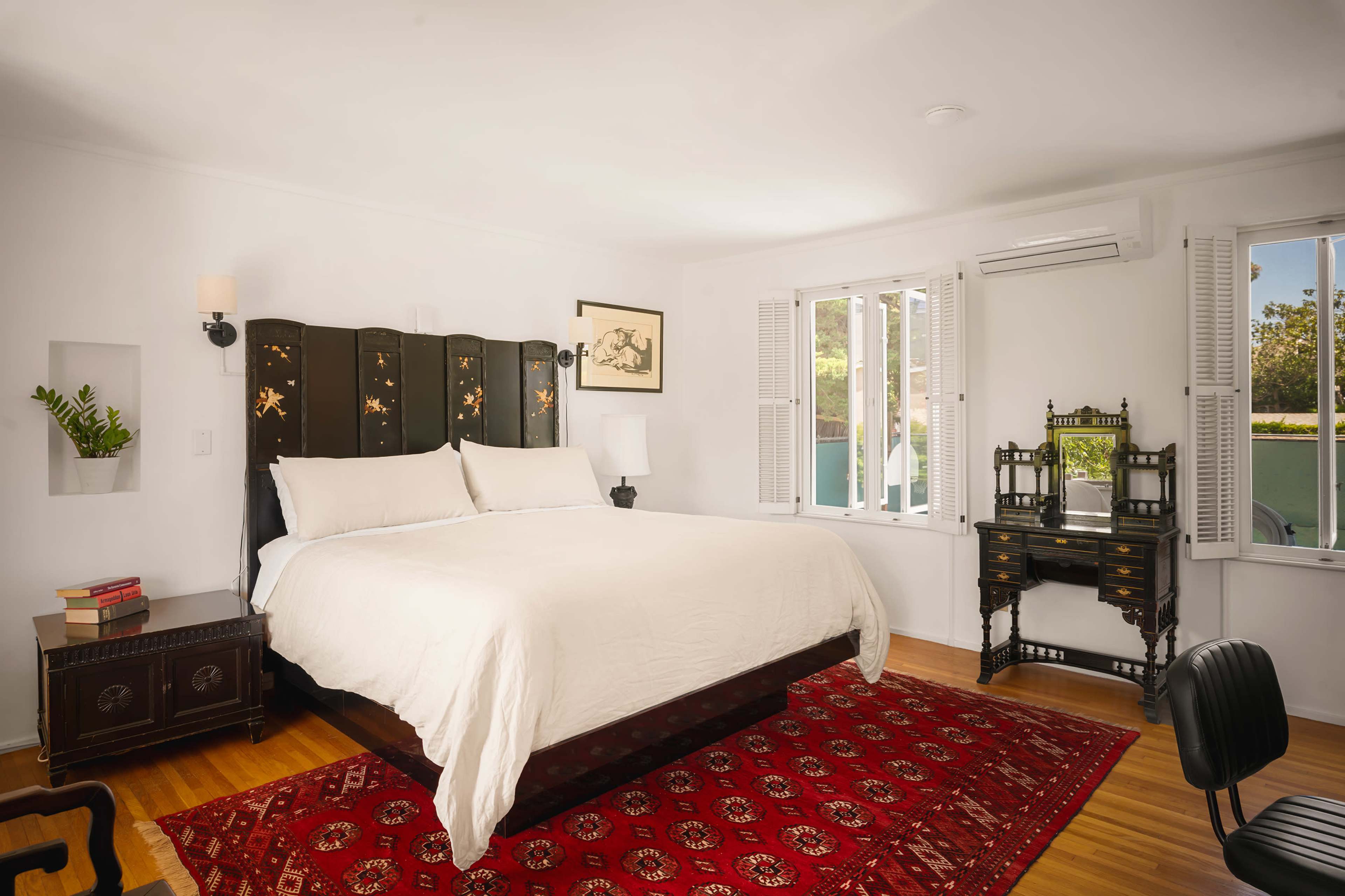 The bedroom features a large bed with a dark wooden headboard, a decorative desk in the corner, and large windows allowing natural light to enter.