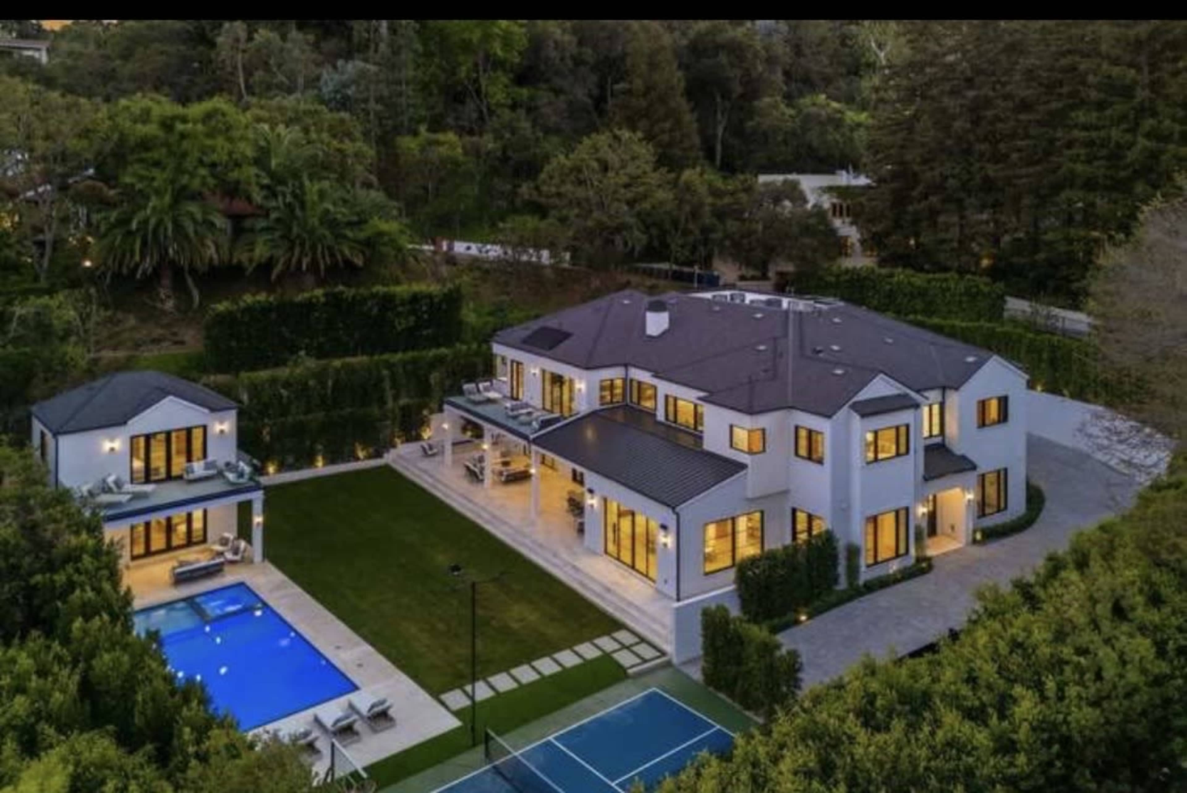 The image shows a large modern house with multiple windows, a swimming pool, a tennis court, and surrounding greenery.