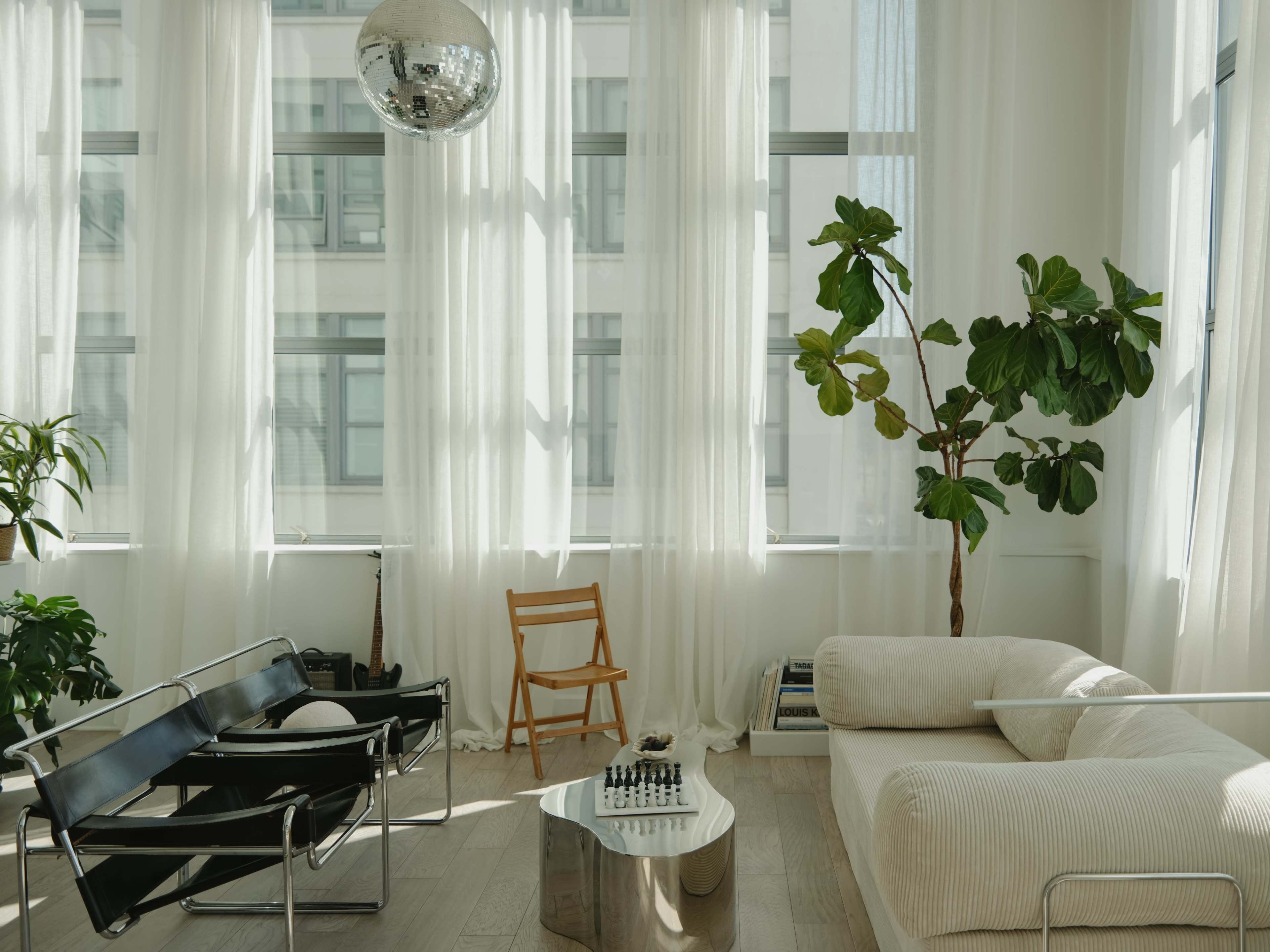 The scene features a modern living room with large windows, sheer curtains, a potted plant, a chess set on a metallic coffee table, and a disco ball hanging from the ceiling.
