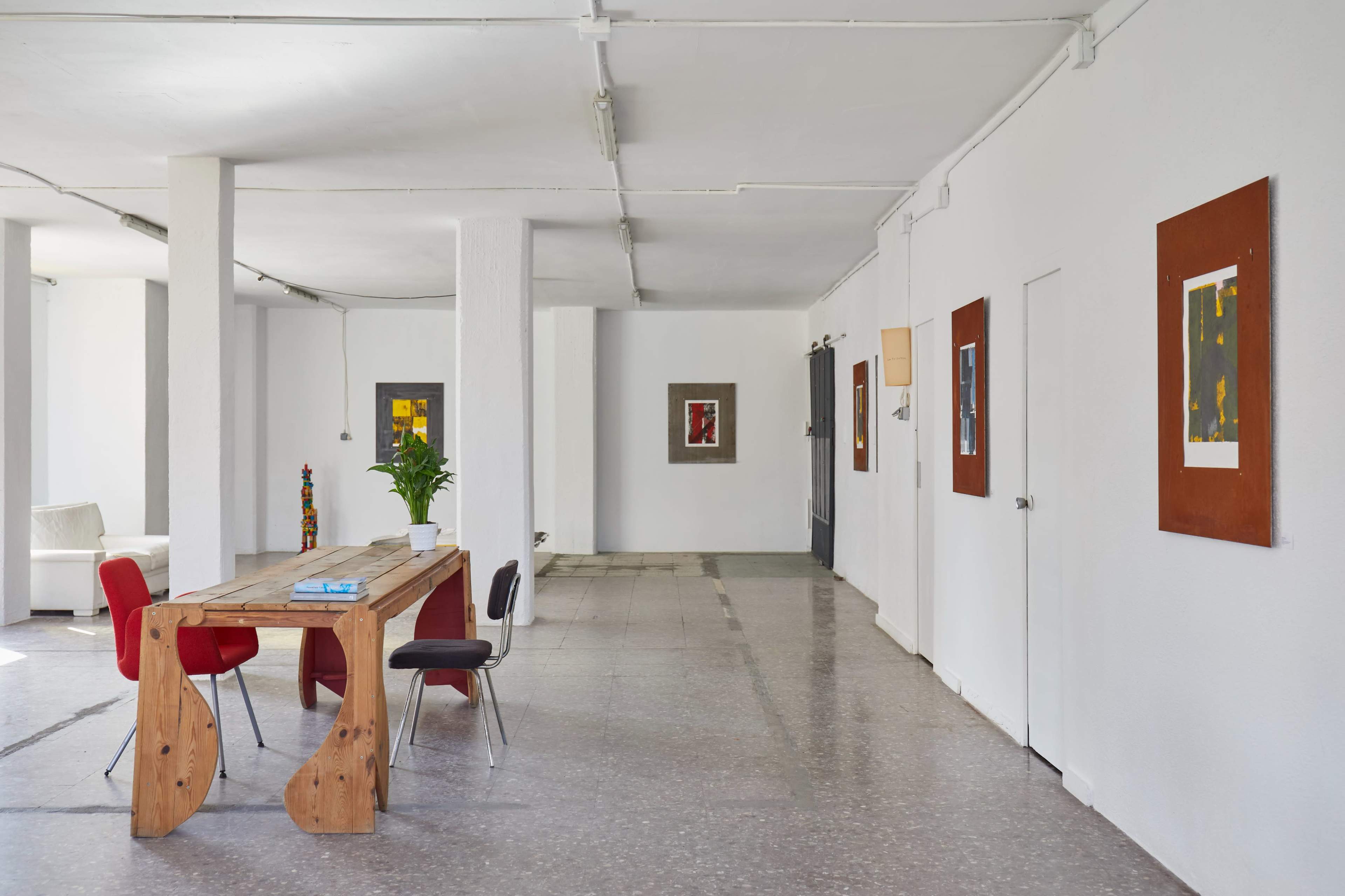 A spacious room with a wooden table and four chairs, illuminated by natural light, featuring artwork on the white walls.