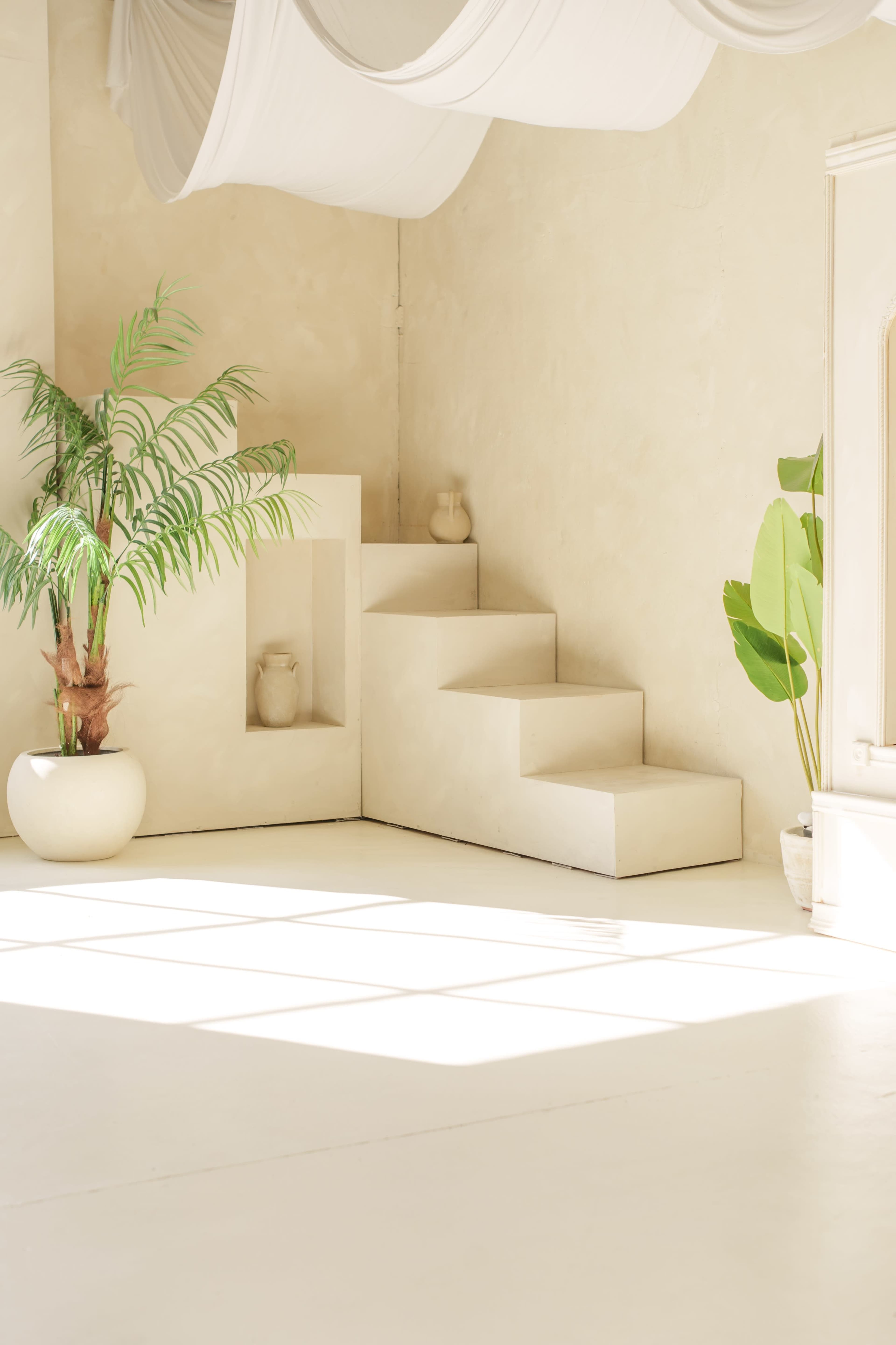 The image shows a minimalist interior space with light-colored walls and a series of white steps leading to a raised platform, accompanied by potted plants.